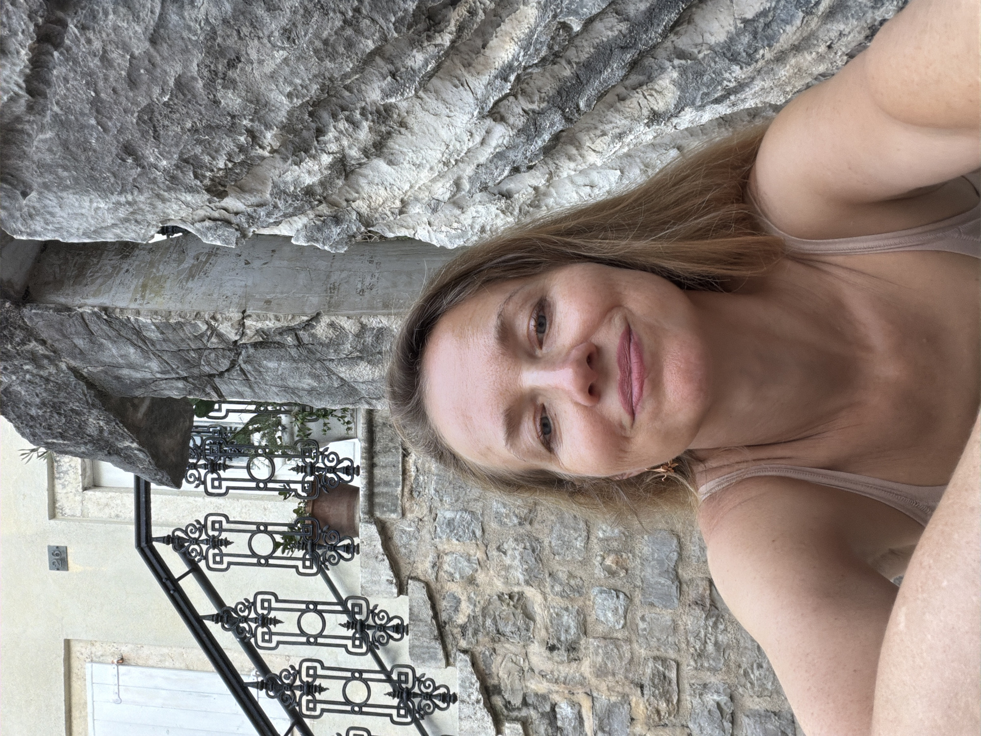 In Budva, Montenegro, a woman with long, blonde hair, wearing a tan tank top, is pictured outdoors. She is positioned in the foreground, looking towards the camera with a slight smile. Behind her, to her left, is a stone wall with a black wrought-iron railing leading up a staircase. A potted plant is visible on the stairs. To her right, a large, textured rock formation dominates the background. The overall environment appears to be an outdoor setting with architectural and natural elements.