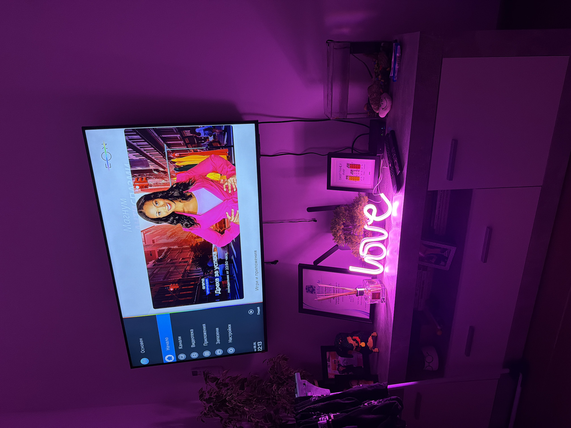 The image depicts a dimly lit living room, illuminated by bright pink ambient lighting. A large television screen displays a menu with Bulgarian text, featuring a woman in a pink blazer. Below the television, a grey and white media console holds various items: a neon pink "love" sign, a diffuser bottle, framed pictures, a small fish tank, and remote controls. The overall scene suggests a modern, personalized living space. The scene is located in Plovdiv, Bulgaria.