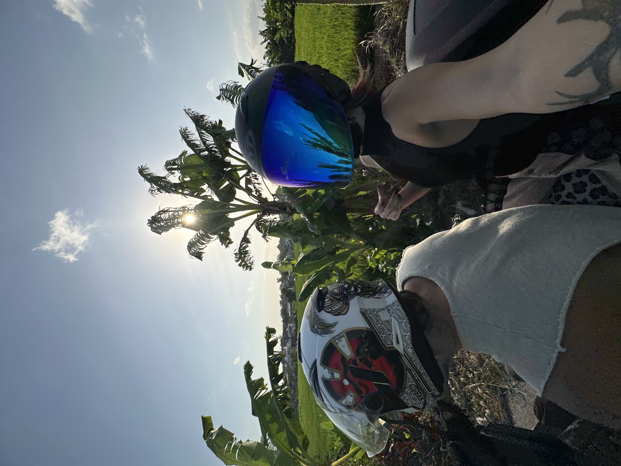 Two individuals wearing helmets are seated on a vehicle, facing away from the camera, with a rice paddy field and lush greenery in the background. The sun is setting or rising, casting a warm glow and visible through the leaves of banana trees. The person on the left wears a white helmet with intricate red and black graphics. The person on the right wears a black helmet with a reflective blue visor. Both individuals are dressed casually. The scene appears to be in a rural area, possibly on the outskirts of North Kuta, Indonesia, given the agricultural landscape and tropical vegetation. The lighting suggests it is late afternoon or early morning.