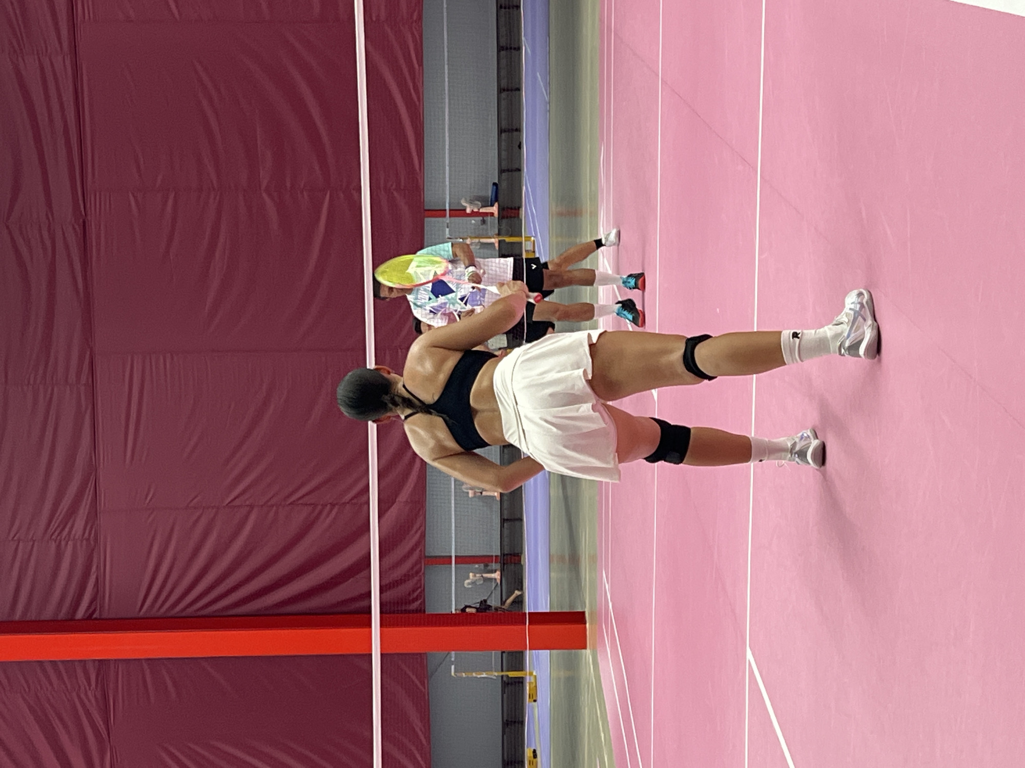 A group of people are playing badminton on a pink court. The main subject, a woman in a black sports bra and white skirt, is in the foreground. She is facing away from the camera and is in a ready stance, with her right leg bent and her left leg extended. She is wearing white socks and athletic shoes, and black knee pads. In the background, several other individuals are visible on the court, some holding badminton rackets. The court is marked with white lines. The walls of the indoor facility are covered with maroon fabric. A red beam runs horizontally across the lower part of the image. The lighting appears to be artificial, suggesting an indoor setting. The scene is likely taking place during the day, as indicated by the bright lighting. The location is Ko Samui, Thailand.