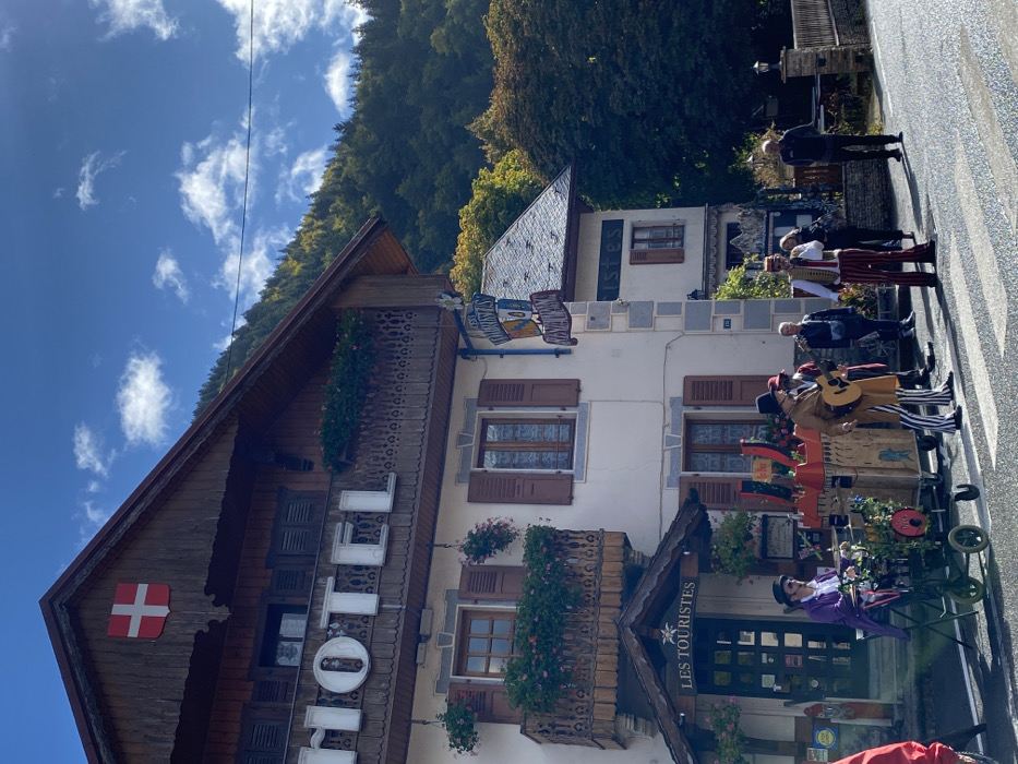 The image depicts a street scene in Abondance, France, featuring the Hôtel Les Touristes on a sunny day. The prominent building, identified by large "HOTEL" lettering and a hanging sign reading "Les Touristes HÔTEL & RESTAURANT," is a multi-story structure with a brown wooden gable roof adorned with a red shield bearing a white cross (Swiss flag emblem). Balconies with decorative wooden railings and flower boxes containing pink flowers are visible on the upper levels. The lower facade is light-colored with stone corner accents and windows with dark brown shutters. An awning over an entrance also reads "Les Touristès."
On the asphalt road in front of the hotel, a group of five individuals is present, including three musicians and two onlookers, positioned near a pedestrian crossing. One musician on the left wears a purple long coat and a black top hat, operating an organ grinder on a two-wheeled cart. Another musician, dressed in a brown long coat, black and white striped trousers, and a black top hat, plays an acoustic guitar next to a large, elaborate wooden musical prop with red accents. A third musician, wearing a red and white striped costume with a vest and a red hat, holds a microphone. Two individuals observe the performance from behind the musicians on the right side of the road.
In the background, a dense forest covers a mountain under a blue sky with scattered white clouds. To the immediate right of the main hotel building, an adjacent structure with a grey sloped roof and similar architectural features is visible. A low stone wall lines the right side of the road.