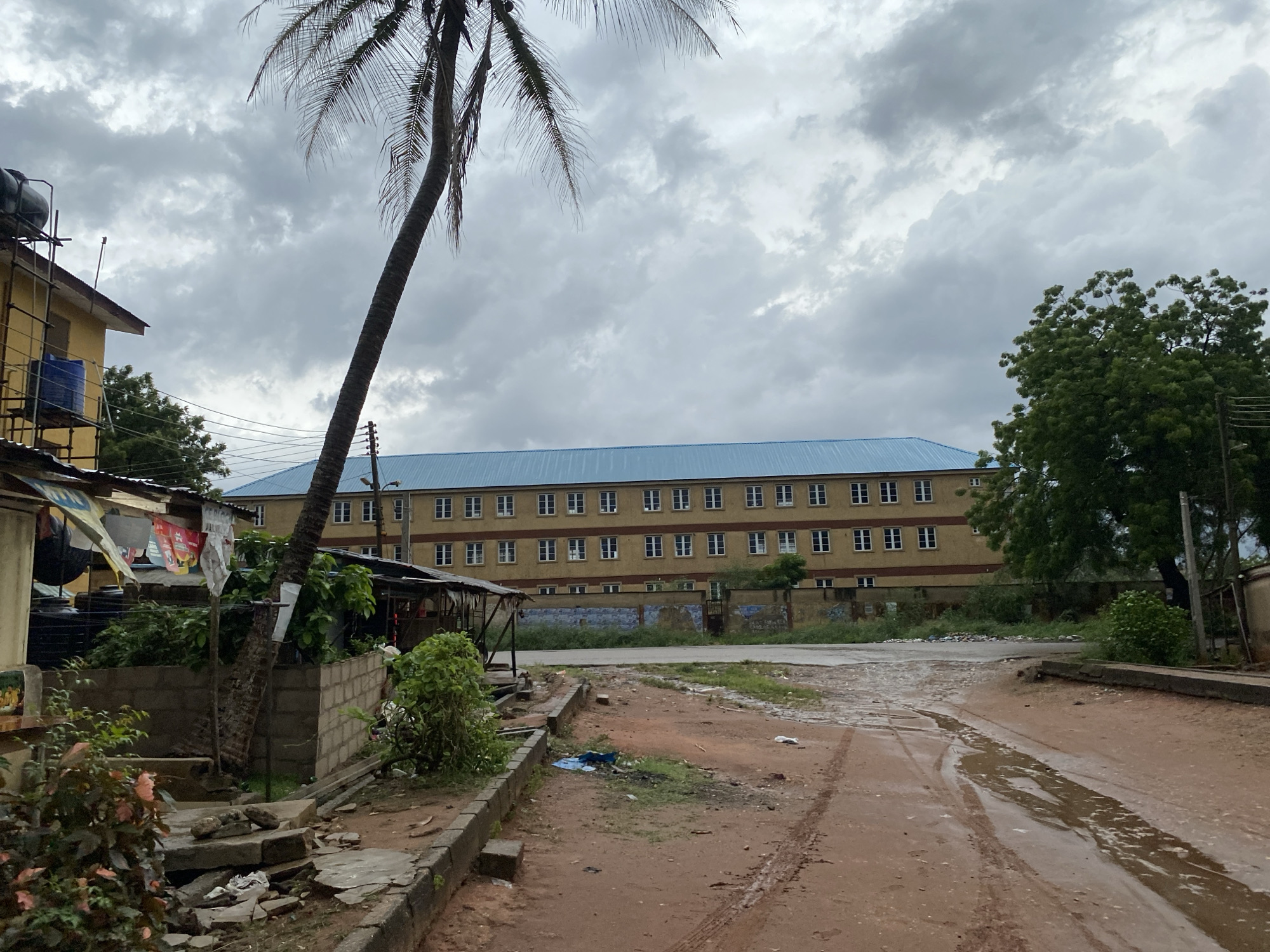 The image depicts a street scene in Lagos, Nigeria, under a cloudy sky. The road is unpaved, with visible tire tracks and puddles of water. To the left, a yellow building with multiple stories is partly visible, with some structures like tanks and scaffolding on its exterior. A palm tree leans towards the left side of the frame. Next to the building is a small roadside stall with hanging banners. A concrete curb separates the stall area from the road. In the background is a long, light-brown building with a blue roof and rows of windows. Vegetation grows in front of the building. To the right of the building is a tree with dense green foliage.