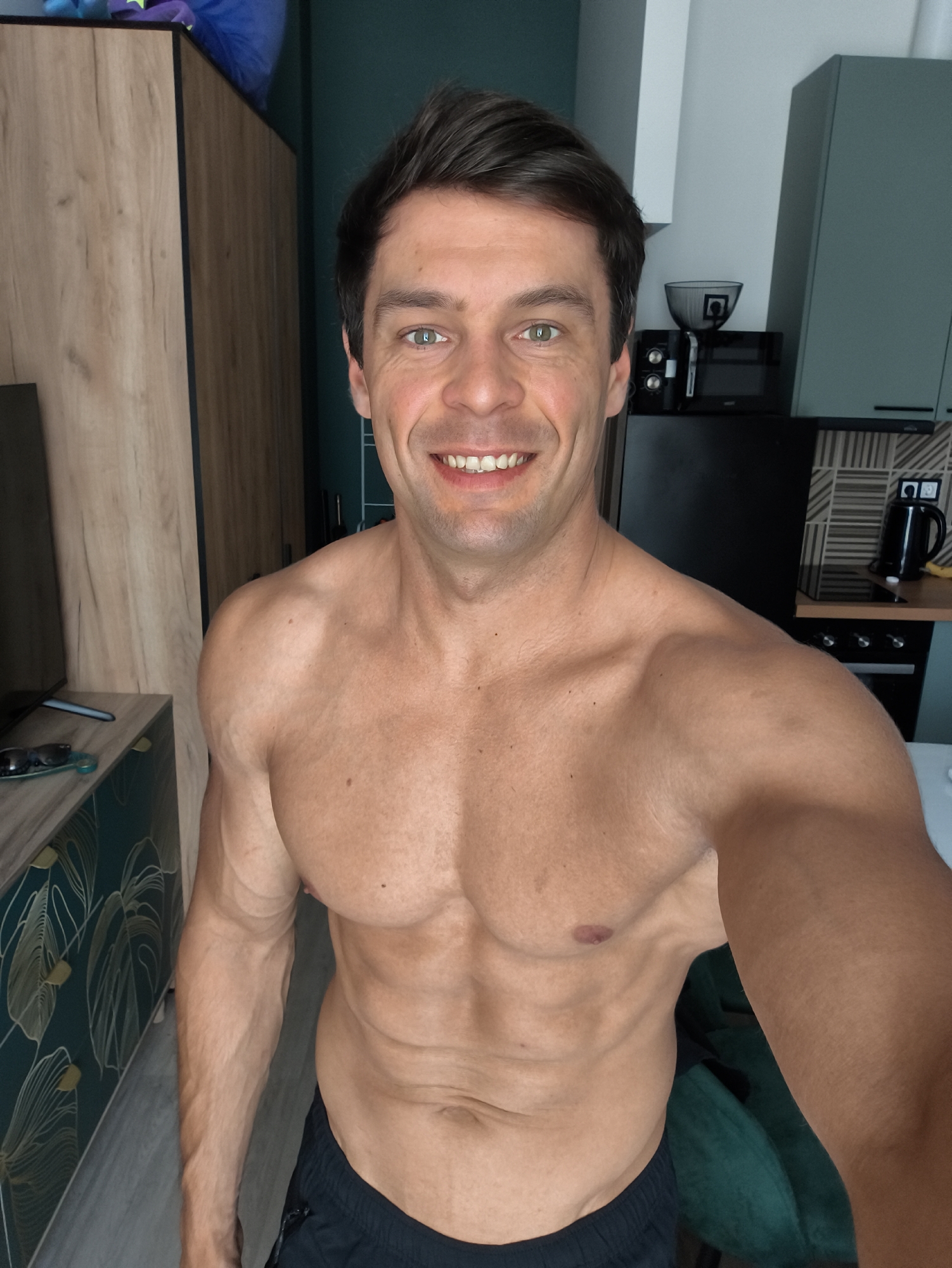 A shirtless man with green eyes and dark hair smiles at the camera. He is standing in what appears to be a kitchen or living area, with a wooden cabinet to his left and a black refrigerator and kitchen cabinets to his right. The man is wearing black shorts. The image is a selfie taken from chest-level.