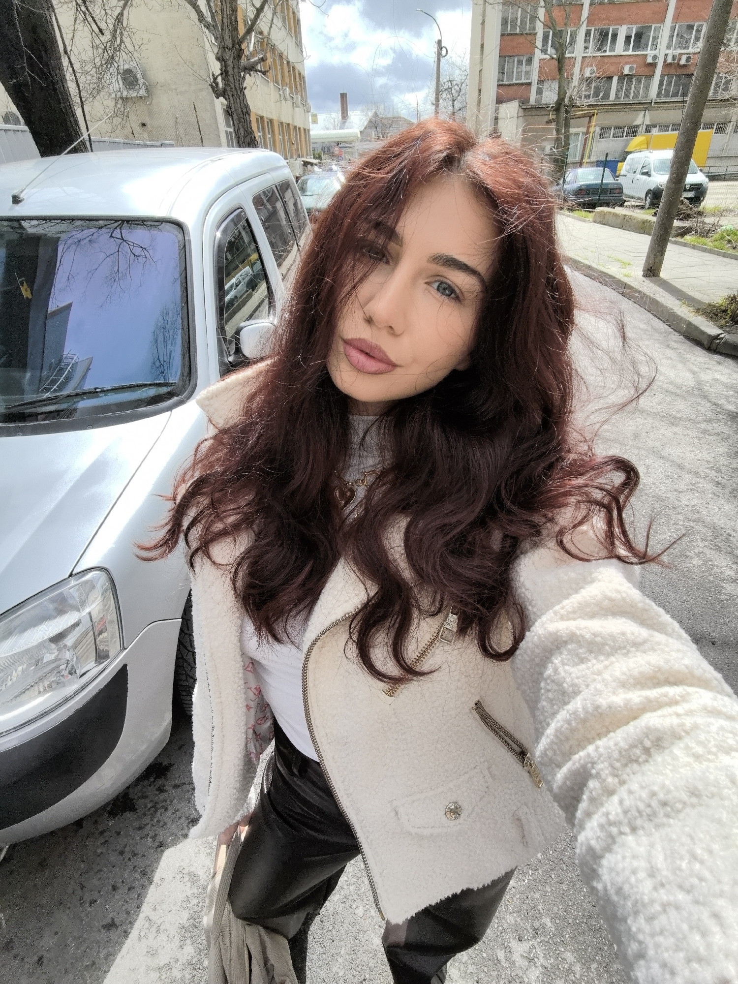 A young adult female is centered in a selfie, looking directly at the camera. She has long, wavy, reddish-brown hair, blue eyes, and pinkish lips. She wears a white turtleneck top, a cream-colored textured jacket with gold-toned zippers, and black leather-like pants. A gold-colored necklace with a heart pendant is visible. She stands on an asphalt surface, likely a street or sidewalk, under direct sunlight, which casts distinct shadows.
To her left, partially visible, is the front section of a silver-colored panel van or minivan, specifically its bumper, headlight, and windshield. The windshield reflects bare tree branches and a blue sky. In the background, an urban street scene unfolds. Several buildings line the street; immediately behind the van is a light-colored multi-story building with multiple windows and an external air conditioning unit, partially obscured by bare deciduous trees. Further down the street, a larger red-brick building with numerous windows and several external air conditioning units is visible. Additional vehicles, including a dark sedan and a white van, are parked along the street. The sky above is partly cloudy. The scene is located on an urban street in Stara Zagora, Bulgaria.