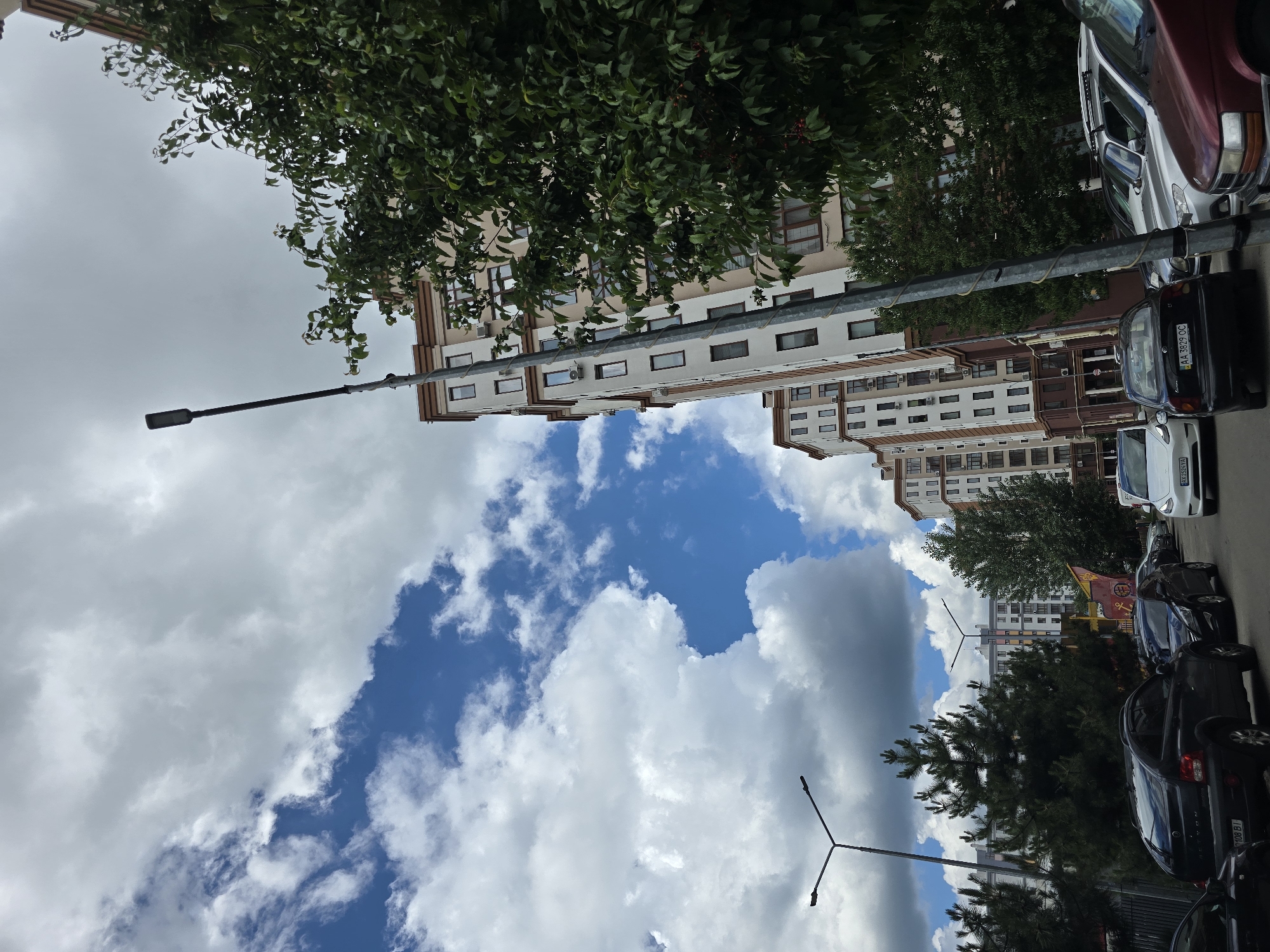 In Petropavlivska Borshchahivka, Ukraine, a street scene unfolds beneath a dramatic sky filled with both bright blue patches and large, billowing clouds. The perspective is angled upwards, emphasizing the verticality of several modern apartment buildings, distinguished by their light-colored facades with accents of brown and beige, and numerous visible windows and air conditioning units. A tall, dark lamppost with a single light fixture stands prominently in the foreground, leaning slightly to the right. Dense green foliage from a large tree partially obscures the right side of the apartment buildings and extends towards the top of the frame. In the lower portion of the image, a row of parked cars lines the street, including a black SUV in the immediate foreground with a partial license plate visible, followed by other vehicles. Another lamppost is visible in the middle ground, and more apartment buildings stretch into the distance, suggesting a residential neighborhood. The overall impression is a contemporary urban environment on a cloudy day.