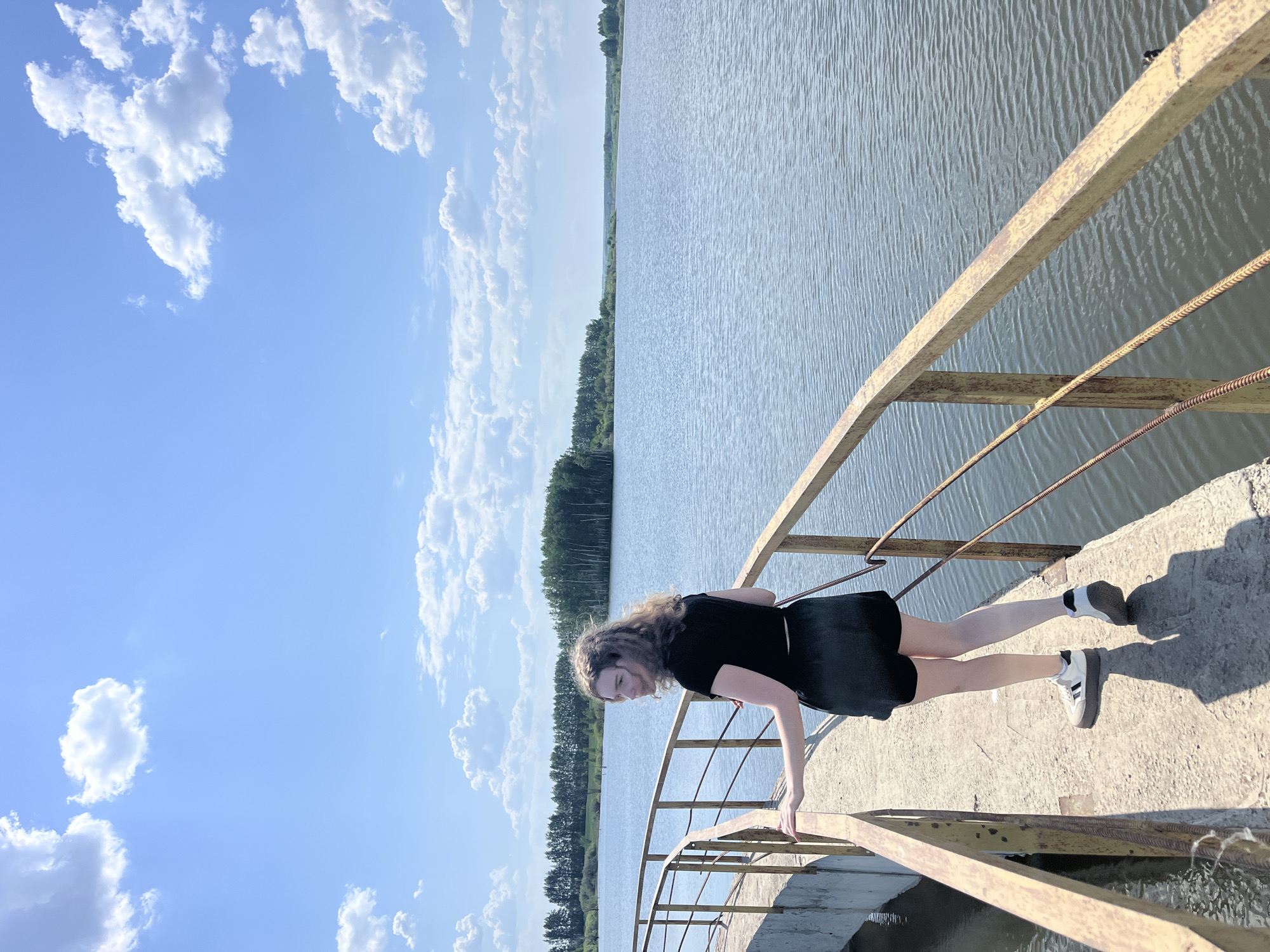 A young person is on a bridge over water, possibly a canal or river. They are wearing a black short-sleeved shirt, a black skirt, and white sneakers with black stripes. The person is leaning forward, with their right hand touching the railing of the bridge. The bridge has a yellow rusted metal railing and a concrete surface. The water is calm with small ripples. Trees line the opposite bank of the water. Above, there is a blue sky with scattered white clouds. The location is Korovino, Russia.