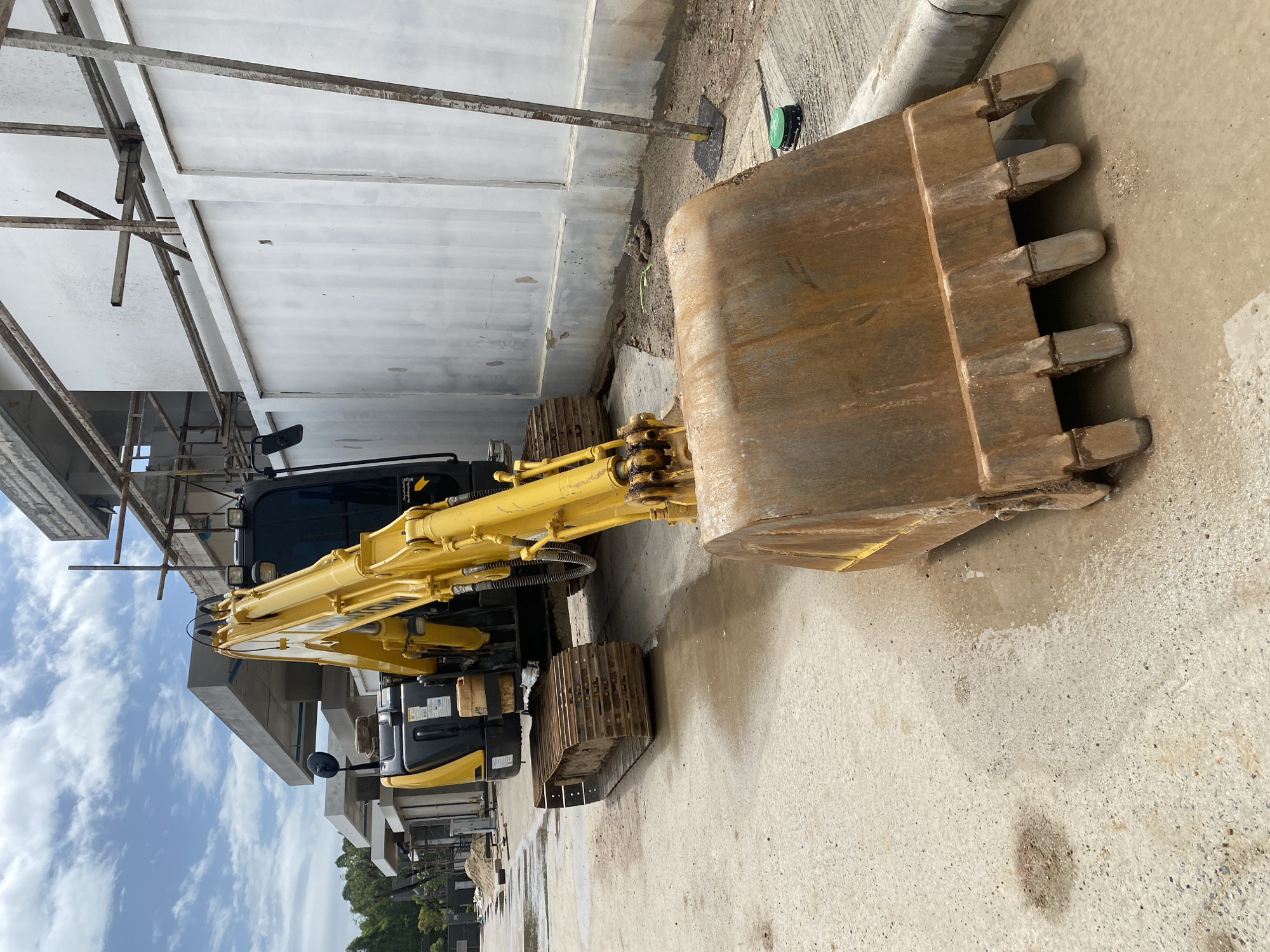 The scene depicts a yellow and black excavator parked on a concrete surface, adjacent to a white building under construction, potentially in Choeng Thale, Thailand. The excavator bucket is prominent in the foreground, exhibiting signs of wear and rust. Scaffolding is visible around the building, suggesting ongoing construction activity. The sky is visible in the background, with scattered clouds. No people are visible in the frame.