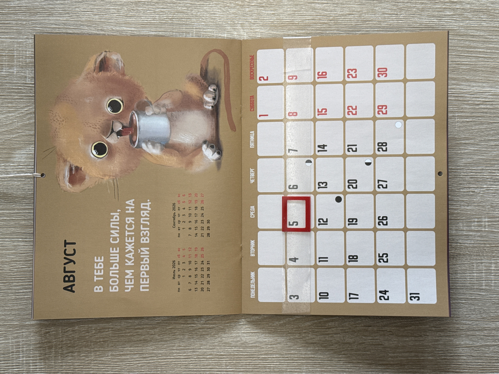 The image is a close-up, eye-level, indoor shot of a calendar page for August, featuring a whimsical illustration of a cartoon lion cub drinking from a mug. The calendar is printed on textured, light brown paper and is mounted on a wall with a light-colored wood grain texture. The top of the page displays the word "АВГУСТ" (August) in large, dark brown lettering, followed by a motivational quote in Russian. Below the quote are mini-calendars for July and September 2026, showing the days of the week and dates. The main calendar grid for August is visible below, with days of the week labeled in Russian (ПОНЕДЕЛЬНИК, ВТОРНИК, СРЕДА, ЧЕТВЕРГ, ПЯТНИЦА, СУББОТА, ВОСКРЕСЕНЬЕ). The number "5" for Wednesday is highlighted with a red square border. Individual dates are displayed in white boxes with rounded corners. Some dates have small circular symbols, indicating lunar phases. The image does not contain any information about the city of Lyubertsy, Russia.