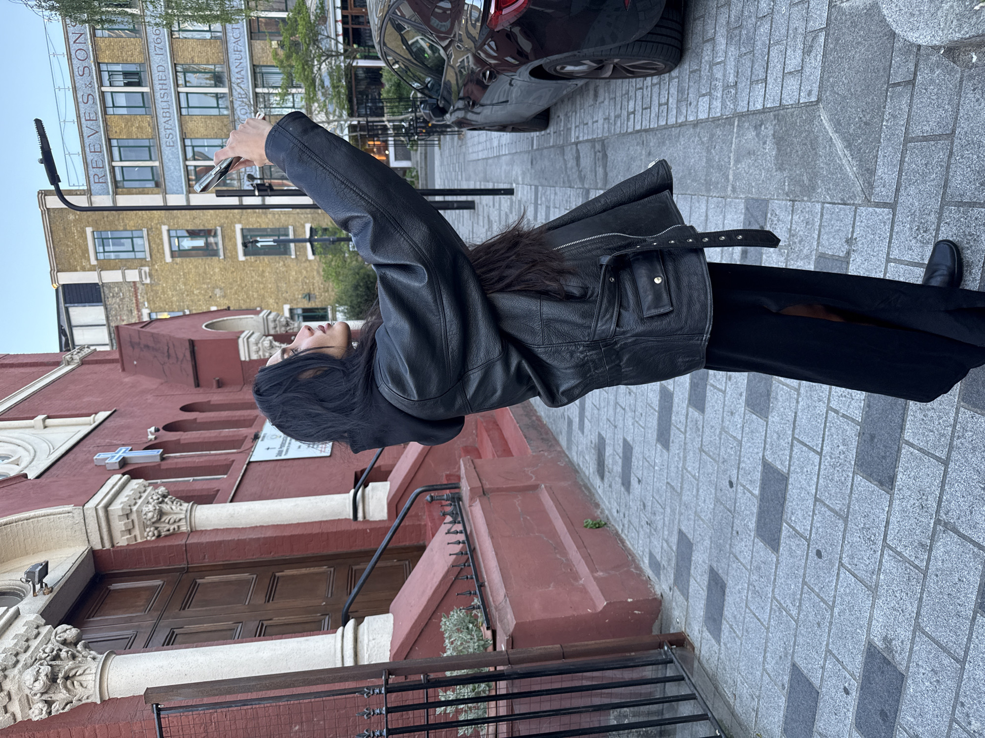 A woman is captured mid-action on an urban street, holding a smartphone high to take a photo or selfie. She is dressed in a black leather jacket with a belted waist, a long black dress or skirt, and black shoes. Her long, dark hair cascades down her back.
The scene features distinctive architecture. To her left, a red-painted building with white ornate columns and large wooden doors, possibly a church, is visible. A blue cross and partially readable text "ST. MICHAEL'S AND ALL ANGELS" are present on its facade. Further in the background, a light brown multi-story building stands out with large windows and signage that clearly reads "REEVES & SONS" and "ESTABLISHED 1766", with "COMB MANUFACTURE" partially visible below. A dark car is parked on the tiled pavement between these structures, and a modern street lamp is also present.
The street is paved with a pattern of light and dark grey rectangular tiles. The time of day appears to be daytime, possibly late afternoon or early evening, under a clear sky. The setting, with its blend of historic buildings and urban elements, suggests a busy city environment, consistent with London, United Kingdom. The activity conveys a casual or tourist-like moment, capturing the surroundings.