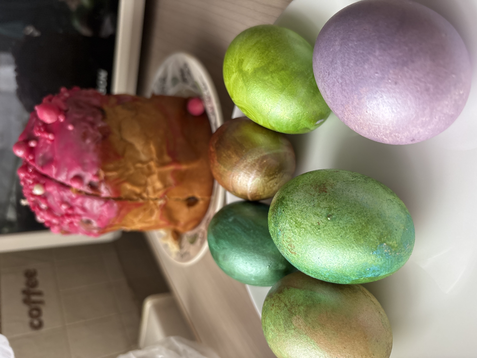 The image shows a white plate with six Easter eggs of varying colors and textures. The colors include green, purple, brown, and blue-green. To the left of the plate is an Easter cake decorated with pink frosting and small white sprinkles. The background includes a "coffee" sign and a blurred-out object. The location in the city of Izhevsk, Russia, is not discernible from the image. There are no visible people, actions, or interactions.