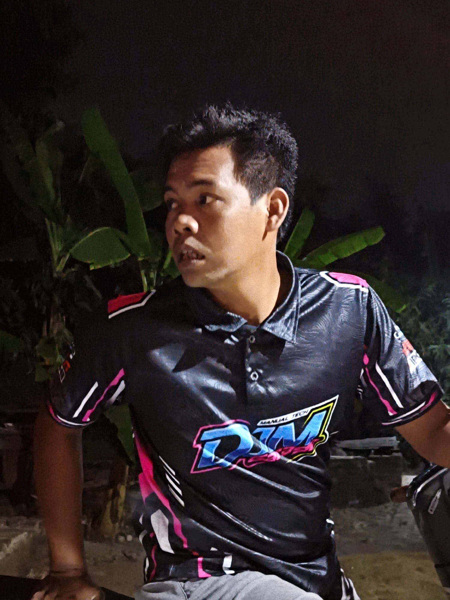 A medium shot shows a man wearing a black polo shirt with colorful graphics and pink and white accents. He has short, dark, curly hair and is looking to his left with his mouth slightly open. The background is dark, with green leaves of a plant visible behind him. The man is sitting, with his arms resting on his lap, and appears to be outdoors at night. The scene is likely in the city of Pengkok, Indonesia, as indicated by the prompt.