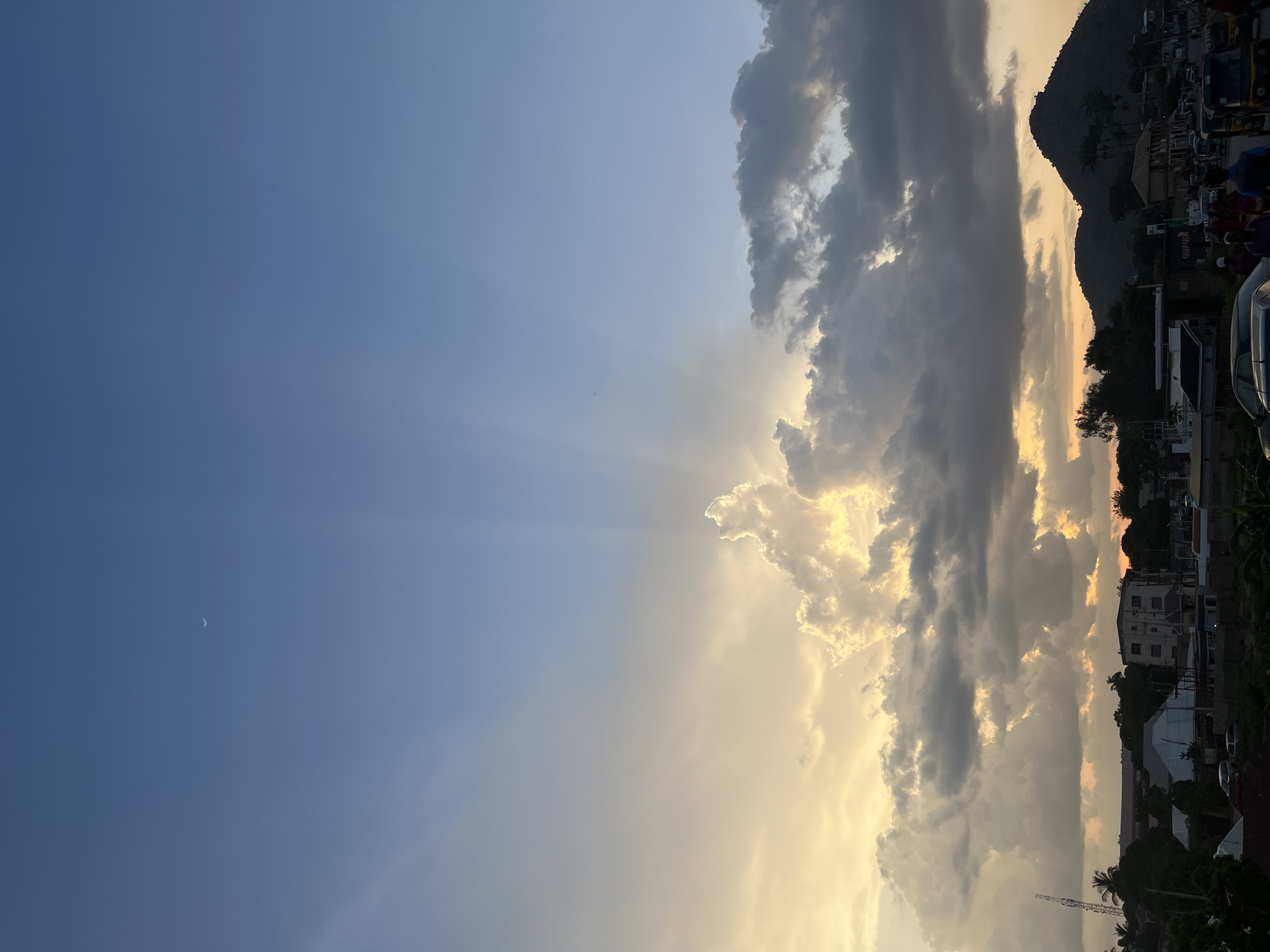 The image depicts a sky over Jos, Nigeria, at what appears to be dusk. The sky is a gradient of deep blue at the top, transitioning to lighter hues of yellow and orange closer to the horizon, suggesting the setting sun. Dramatic clouds, some dark and heavy, others illuminated by the sun's rays, dominate the right side of the frame. The sun's light breaks through the clouds in visible rays, creating a dramatic effect.
In the lower right portion of the image, a silhouetted mountainous landscape is visible, with some trees and buildings at its base. The buildings are a mix of shapes and sizes, suggesting an urban or semi-urban environment. A thin crescent moon is faintly visible in the clear blue portion of the sky on the left. The scene is serene and captures a beautiful natural phenomenon. The overall mood is peaceful and awe-inspiring, highlighting the beauty of the sky during sunset. There are no discernible people or specific objects in focus, with the primary subject being the sky and its atmospheric conditions.