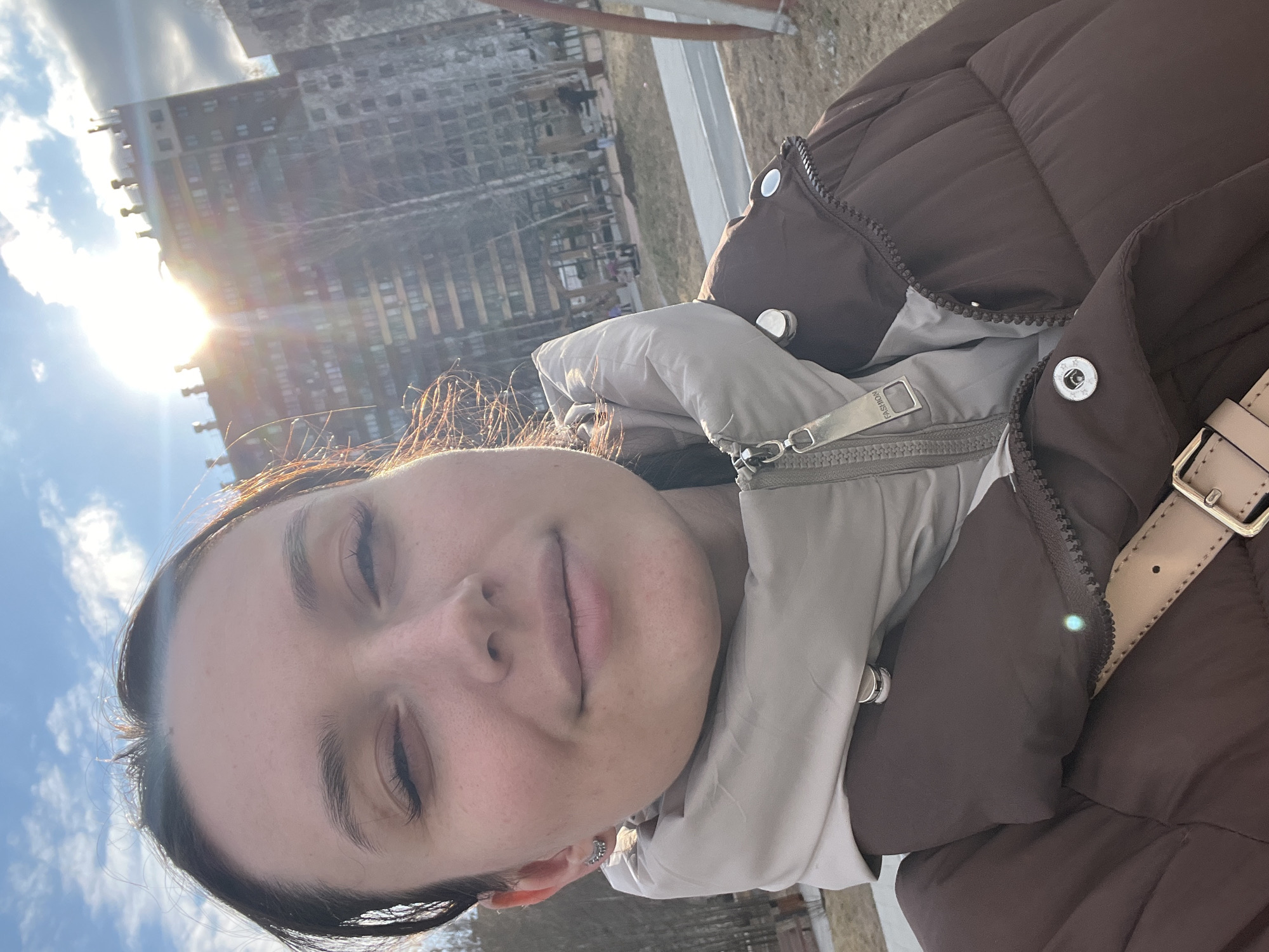 A person with closed eyes takes a selfie outdoors, wearing a brown and beige puffer jacket. In the background is a tall apartment building, partially obscured by the sun and clouds in a blue sky. The scene appears to be located in a city, possibly Tyumen, Russia, based on the architectural style of the building.