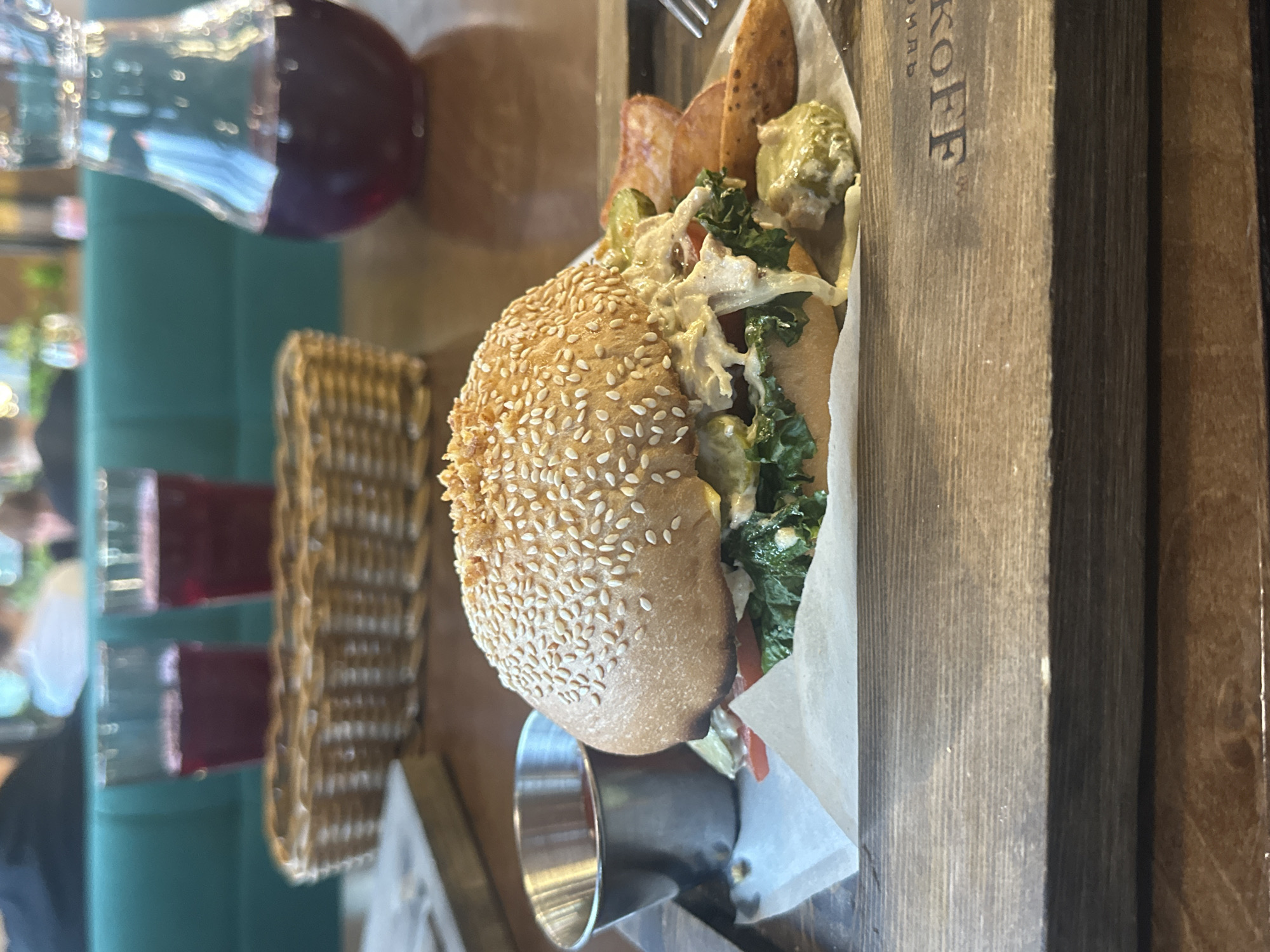 A close-up, eye-level, indoor shot shows a gourmet burger on a wooden serving board. The burger, centered in the frame, has a sesame seed bun, a thick patty, lettuce, tomato, pickles, and a creamy sauce. Next to the burger are potato wedges and a small metal cup filled with red sauce. In the background, two glasses of red liquid and a woven basket are visible. The overall setting appears to be a restaurant in Blagoveshchensk, Russia.