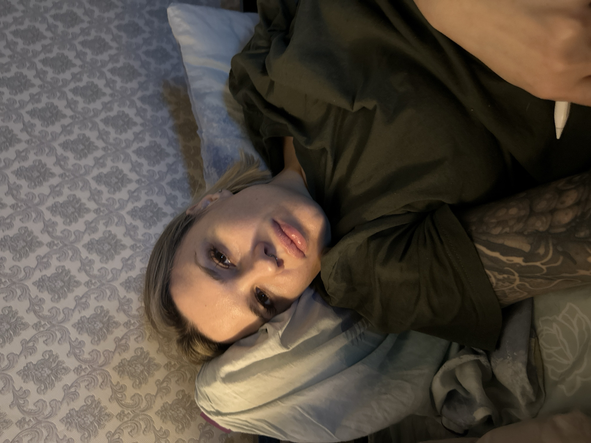 An indoor scene in Rostov-on-Don, Russia, depicts an adult female lying down. Her head rests on a light-colored pillow, and she faces the camera directly. She has blonde, short-to-medium length hair, a septum piercing, and visible facial makeup including false eyelashes. She wears a dark olive-green short-sleeved top. A prominent, dark-inked tattoo featuring intricate, possibly scales or ornamental patterns, covers her left arm. A small, white, cylindrical object is partially visible in her right hand. The background wall is covered with light grey patterned wallpaper displaying a repeating damask-style floral or scroll motif. The ambient lighting is dim.