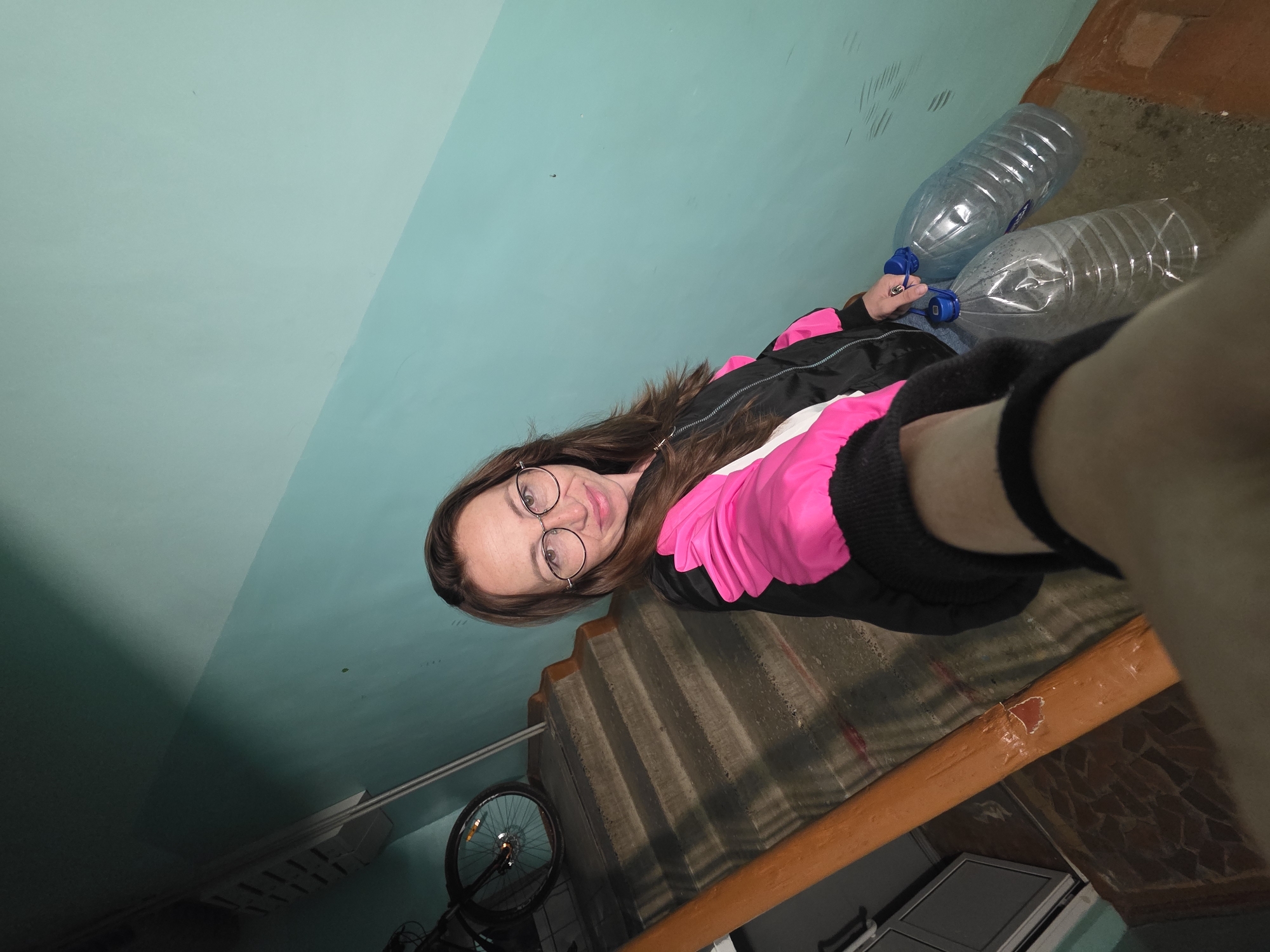A woman is taking a selfie on a staircase in Chaikovsky, Russia. She is wearing glasses, a black jacket with pink accents, and holding two large plastic bottles of water. To the left of the woman, a bicycle is parked on the stairs. The staircase is made of concrete with wooden railings and the walls are painted a light blue-green color. The lighting suggests it may be evening or indoors.
