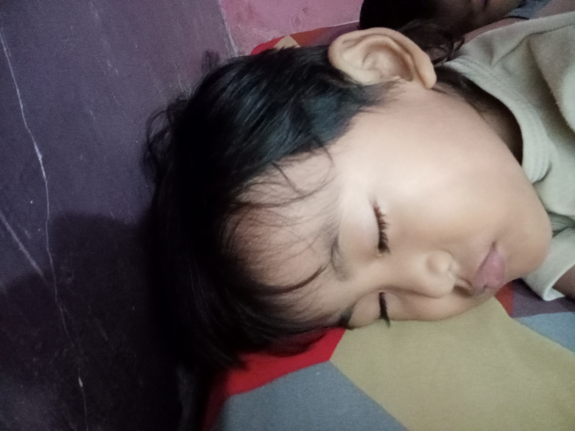 A close-up, overhead, slightly angled shot shows a young child's head resting on a striped blanket. The child is asleep, facing to the right, with their eyes closed and their mouth slightly open. Their dark hair is partially visible, as is their ear. The background consists of a dark purple surface, likely a floor or bed, with some lighter colored markings. To the right, a person's shoulder and arm are visible, wearing a light-colored garment. The scene appears to be indoors. This image could be located in Bloro, Indonesia, but no specific location details are evident from the image itself.