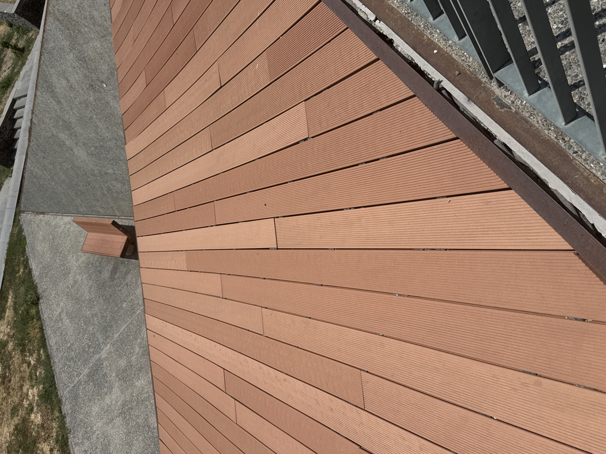 This image shows an outdoor wooden deck with a reddish-brown hue, constructed from parallel planks. The planks have a ridged texture and are laid out in a staggered pattern. The deck appears to be elevated, as indicated by a metal railing system visible along the bottom right edge. Beyond the deck, a pathway made of grey concrete with a textured surface is visible, leading to a grassy area. A small, angled wooden structure, possibly a bench, is situated on the concrete pathway. The scene suggests a public outdoor space, potentially a park or recreational area. The architectural style and materials are consistent with modern urban design, and could be found in various cities. Without further context or specific landmarks, it is not possible to definitively pinpoint this location to Yerevan, Armenia.