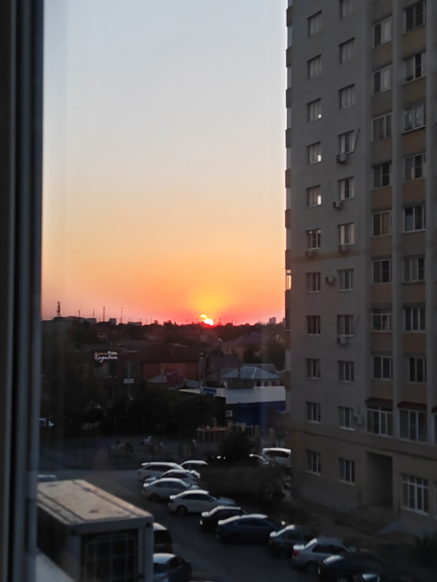This is a vertical, medium-shot, eye-level photograph taken from an elevated perspective, likely an apartment window, looking out over a city street at sunset. The sky is a gradient of soft orange and pink. On the right side of the frame, a tall, light-colored apartment building with numerous windows occupies most of the vertical space. The building has a light gray facade with beige accents around some windows. On the left side of the frame, a dark, vertical element, possibly a window frame or part of a balcony, partially obscures the view.
Below the sky and the apartment building, a cityscape unfolds. There are several smaller buildings, some with dark roofs and others with lighter, modern structures. A sign with stylized white lettering is visible on one of the buildings. The street below is lined with parked cars, including several white sedans and SUVs, and a few darker colored cars. Trees and other greenery are scattered throughout the scene. The sun is setting on the horizon, casting a warm glow over the city. The overall impression is one of a quiet urban evening. The scene is consistent with an apartment building overlooking a street in the city of Bataysk, Russia.