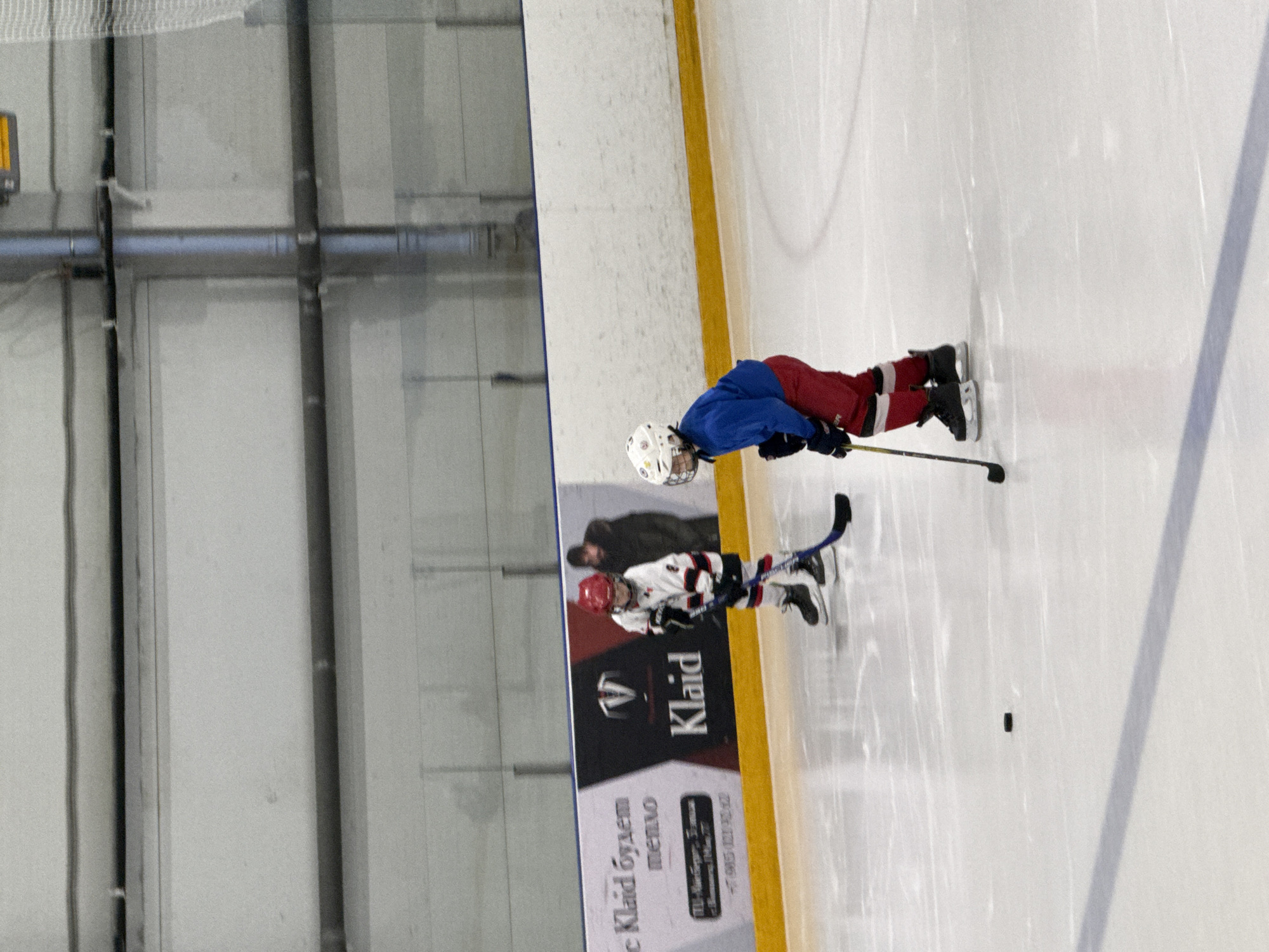 The image shows two young children playing ice hockey on an indoor ice rink. The child in the foreground is wearing a red hockey jersey, blue pants, and a white helmet. They are holding a hockey stick and appear to be in motion. Behind this child, another child in a white and red jersey is visible. The background shows the interior of an ice rink, with white walls and a yellow boundary line. There is also a banner with text in Russian. The lighting suggests it is daytime, and the indoor setting implies consistent weather conditions. The children's postures suggest an active game, with one child leaning forward as if skating or about to shoot. The overall scene is one of youthful recreation in a sports environment. Visible text on the banner includes "Klaid" and other Cyrillic script, suggesting a local business or event advertisement. The location is Votkinsk, Russia.