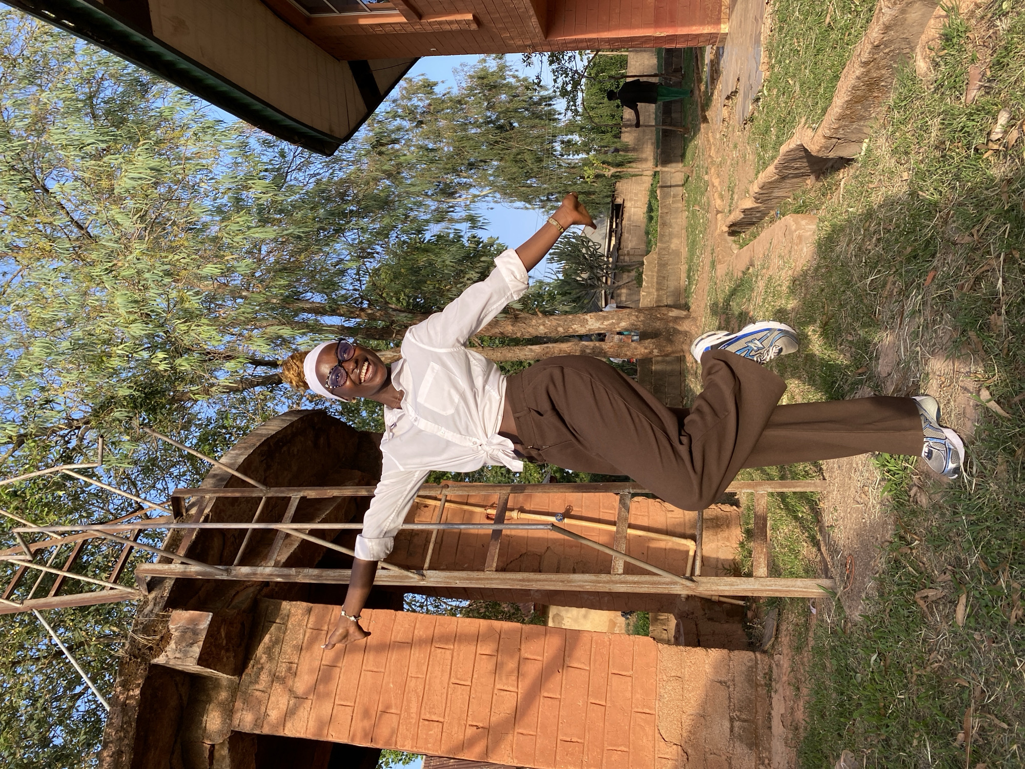 A young, dark-skinned woman with round glasses and a white headscarf is posing outdoors in Vom, Nigeria. She's wearing a white button-down shirt knotted at the waist and brown trousers. She is positioned horizontally in the frame, with her arms outstretched. Her right hand is raised in a peace sign. She is wearing blue and silver sneakers. She appears to be standing on an old, weathered brick and metal structure with trees and grass surrounding her. There is a low brick wall and steps in the background, alongside some trees and another person standing further away. The sky is visible through the trees.