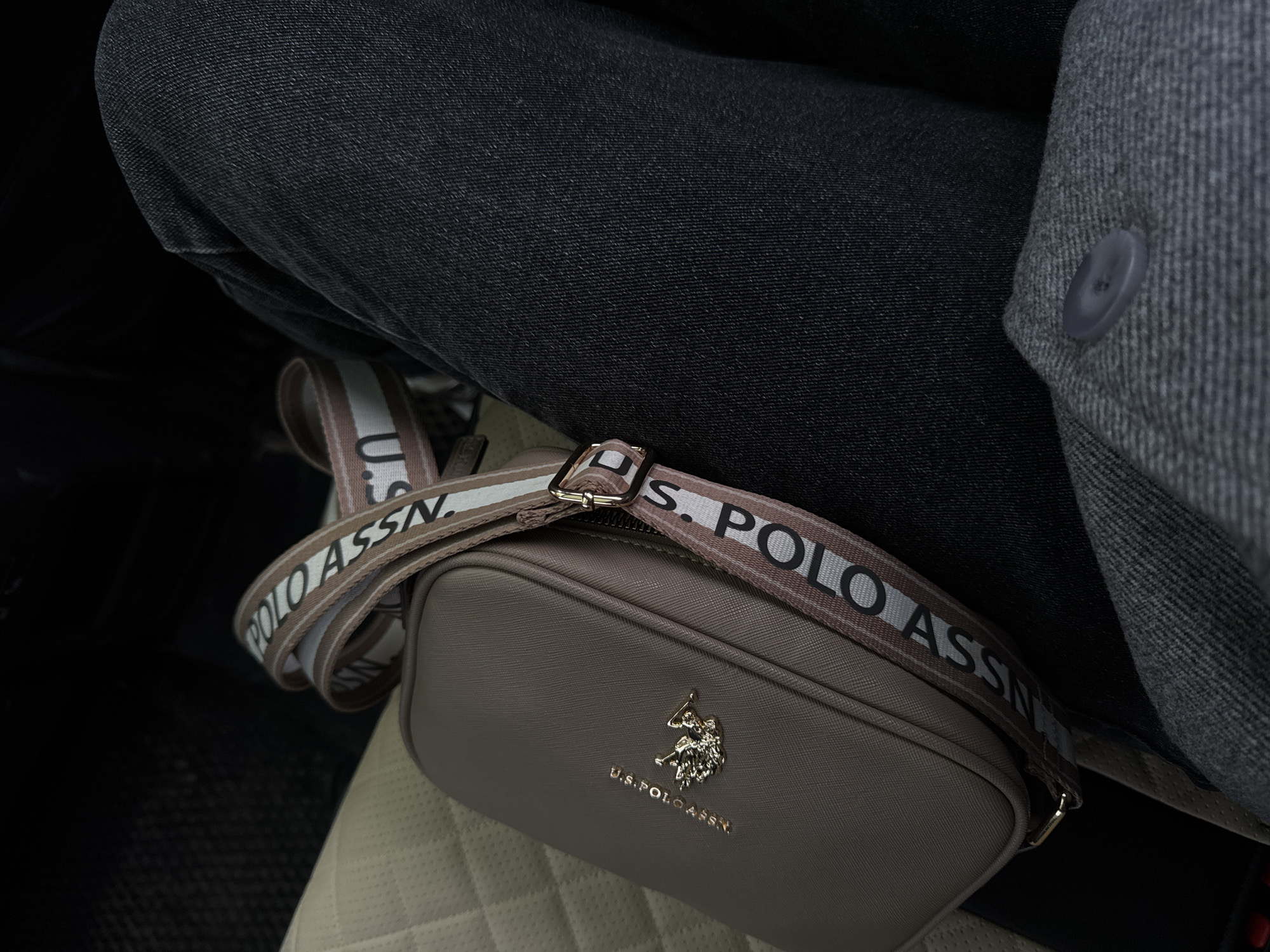 The image depicts a person sitting in a vehicle. The person is wearing dark denim jeans and a gray coat with a button. A U.S. Polo Assn. crossbody bag rests on the person's lap. The environment appears to be the interior of a car with cream-colored, quilted leather seats. There is no indication of the city of Perm, Russia.