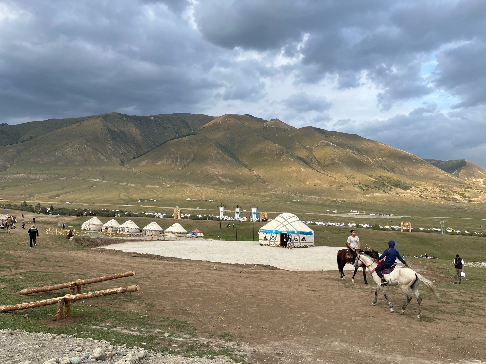 In Grigor'yevka, Kyrgyzstan, the image captures a wide shot of a semi-arid landscape with rolling hills in the background under a cloudy sky. In the foreground, a dirt path leads to a clearing with several yurts and a few people. Two individuals are riding horses, one on a dark horse and the other on a white horse, moving away from the viewer. A person in a dark jacket walks away from the camera on the left side of the clearing. Several yurts are arranged in a semi-circle on the right side of the clearing, with some people visible near the entrance of the yurts. In the background, a vast field dotted with more yurts and tents stretches towards the hills, with some vehicles and figures sparsely scattered across the landscape. The environment appears to be an outdoor event or encampment.