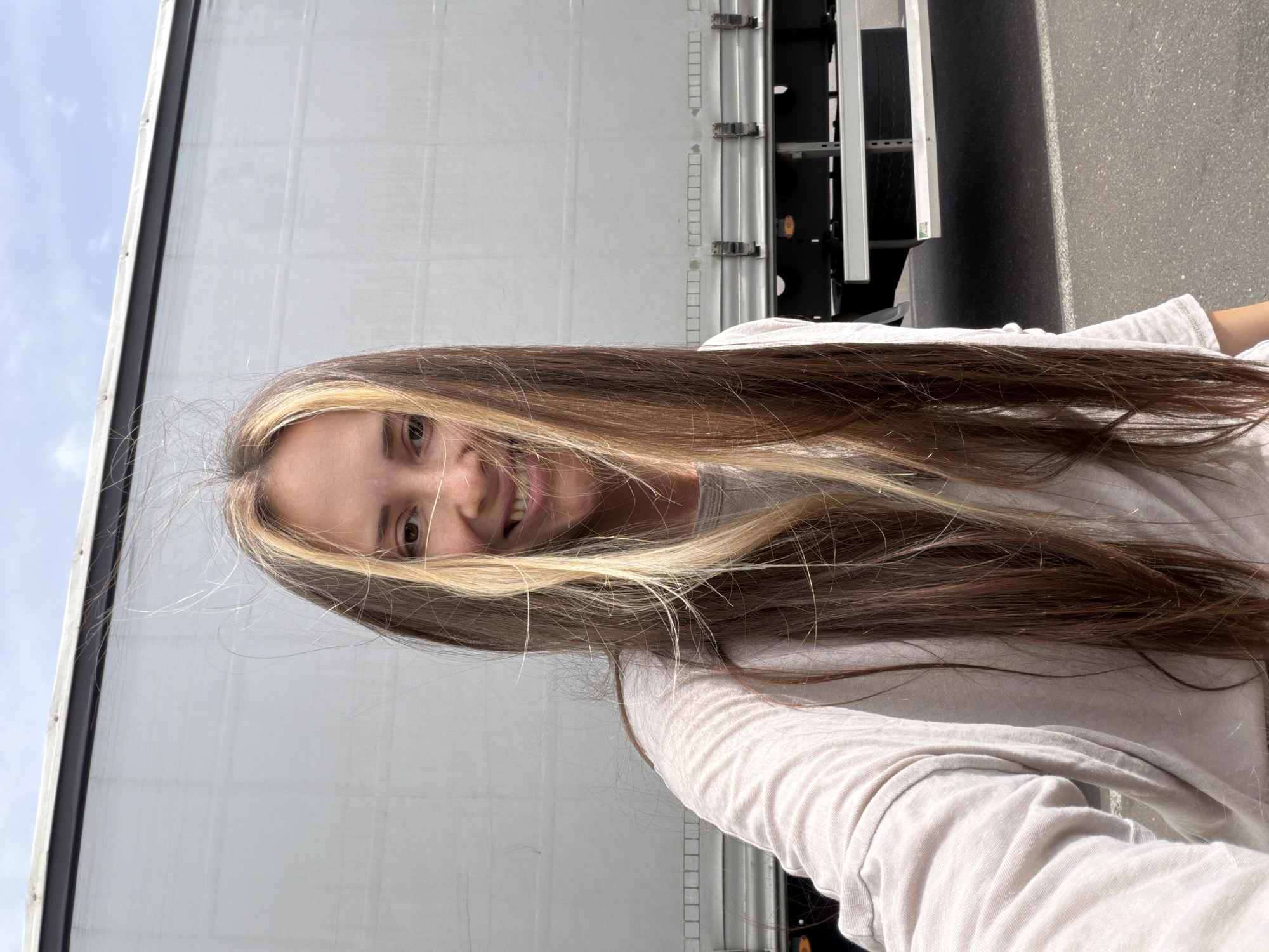 A woman is standing next to a truck. The location is the city of Sluchainyi, Russia. The woman has long brown hair with blonde highlights and is wearing a light-colored long-sleeved shirt. She is smiling. The truck is white with black components. The background includes a street or parking area and a cloudy sky.