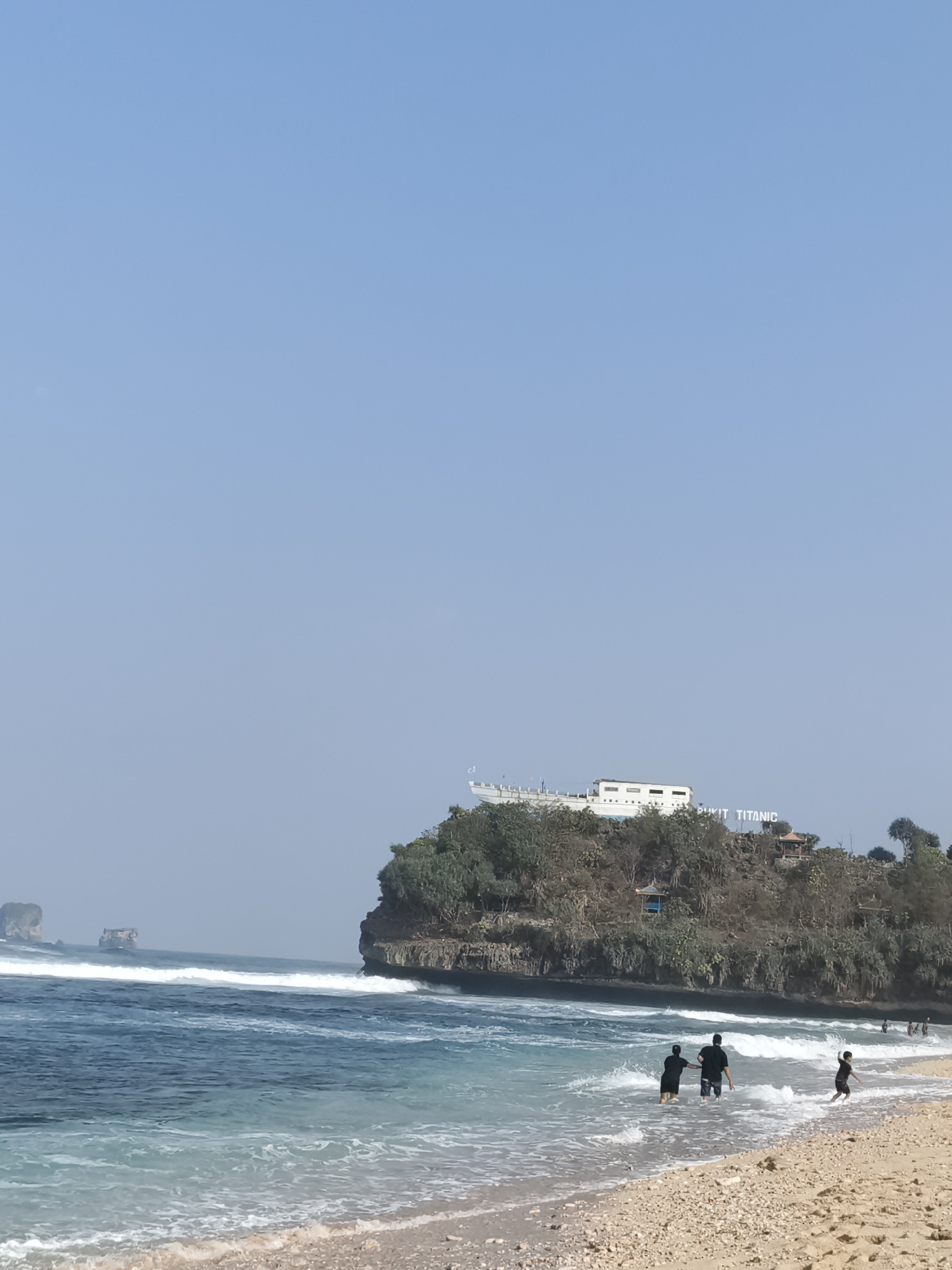In the city of Unknown, Indonesia, a serene beach scene unfolds under a clear blue sky. The turquoise ocean laps at the shore, with gentle waves breaking and creating a foamy white edge. A sandy beach, interspersed with pebbles, stretches into the foreground.
In the shallow water, a group of people are enjoying the day. Two adults, a man and a woman, stand hand-in-hand, seemingly wading through the water. To their right, a young child runs towards them. Further out in the ocean, several other figures are visible, some appearing to be swimming or playing in the waves.
Dominating the background is a dramatic cliff face, densely covered with lush green trees. Atop the cliff, a striking white structure resembling a ship's hull stands prominently, with the words "PUKIT TITANIC" visible behind it. Small, dark-roofed gazebos or small structures are also scattered amongst the trees on the cliff. In the distance, two large rock formations rise out of the ocean, adding to the scenic beauty of the coastline. The overall atmosphere is one of tranquility and leisure.