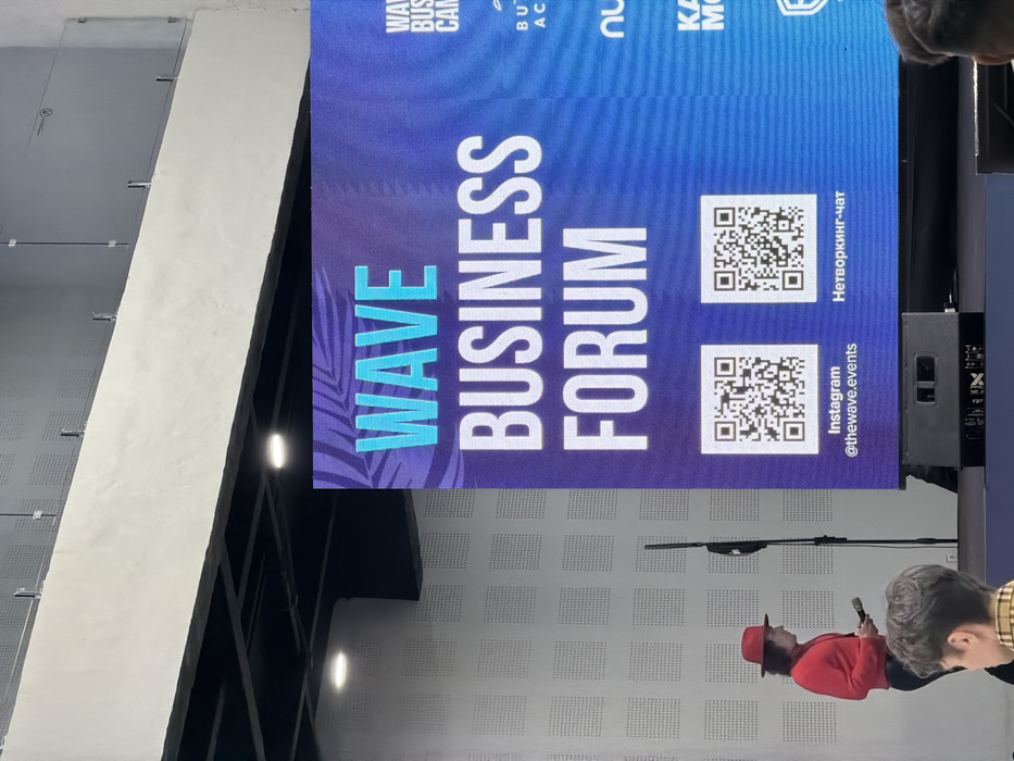 A banner advertises "WAVE BUSINESS FORUM" with text in a vertical arrangement. The background features blue and purple hues with a palm leaf design. Two QR codes are displayed, one linking to Instagram @thewave.events and the other to a "Networking-chat." Parts of the words "Wave Business Camp," "But," "Ac," "Nu," and "Ka" from the upper banner are barely visible. A person is facing away from the camera in front of the banner.