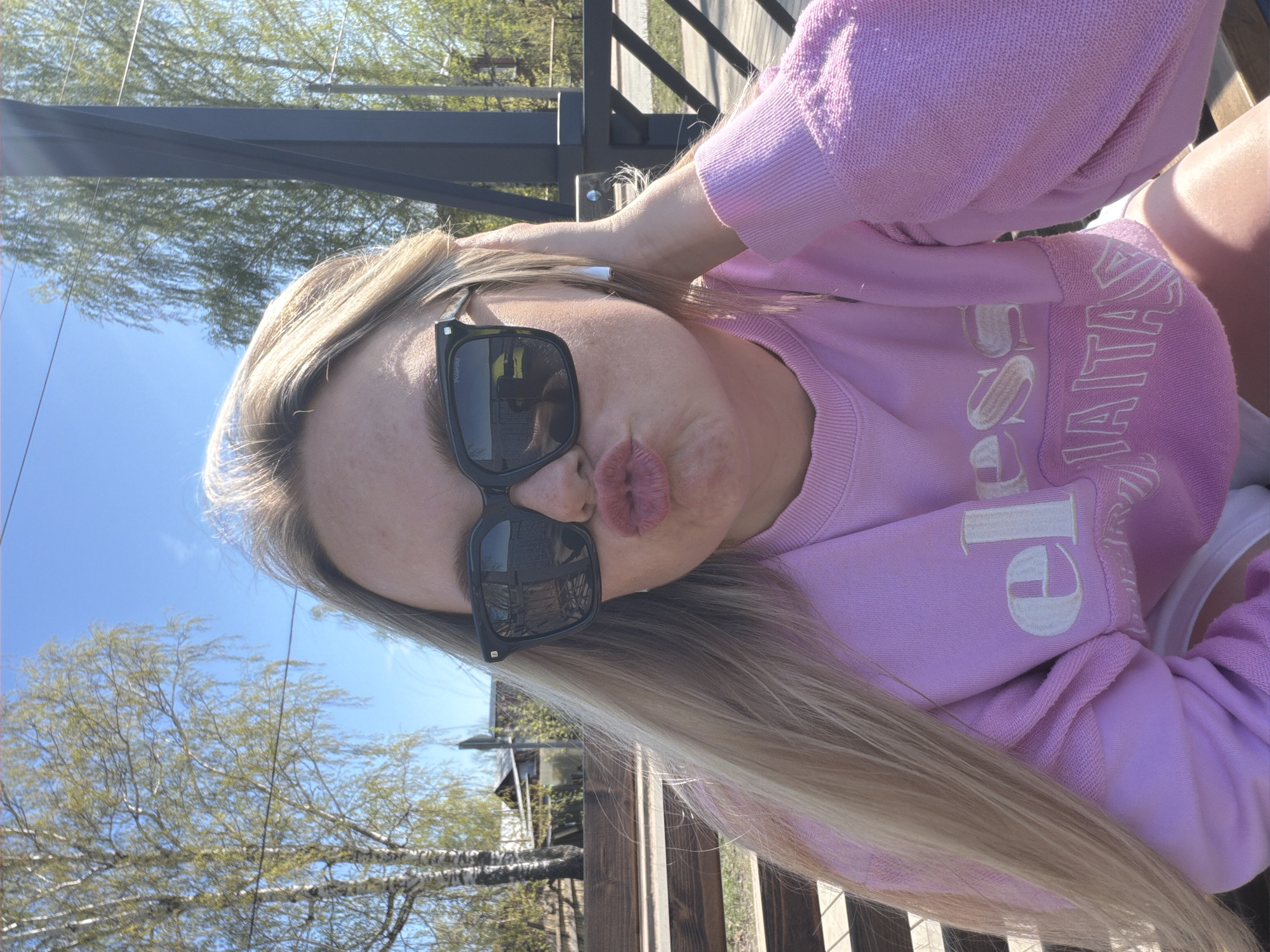 A woman is posing for a selfie outdoors. She has long blonde hair, is wearing black sunglasses and a light pink sweatshirt with a white logo. She is pursing her lips. The background consists of a blue sky, trees, and a wooden fence. The photo appears to be taken near a building. There is no specific identifying feature indicating that this location is in Votkinsk, Russia.