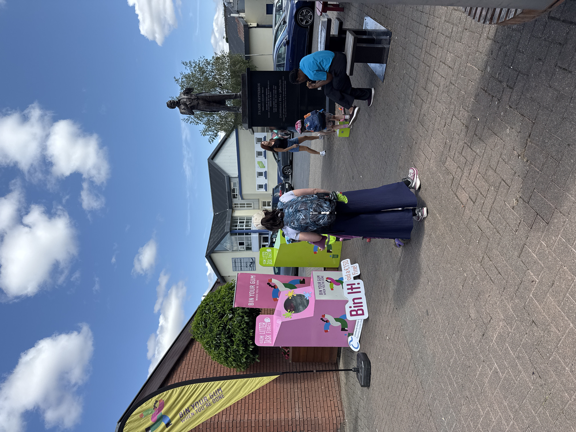 The image depicts an outdoor scene in Clonmel, Ireland, near the Frank Patterson statue. In the foreground, a pink gum bin with cartoonish figures is set up, featuring the text "BIN YOUR GUM WHEN YOU'RE DONE" and "Bin It!". A woman wearing a backpack and wide-leg pants stands in front of the bin. To the right, another gum bin, this one green, is visible. Further back, several people are interacting with the environment: a woman in a black top and denim shorts stands near the bins, a child with a backpack kneels near the green bin, and two adults are seated at a picnic table with a statue of Frank Patterson behind them. The statue is on a plinth with an inscription. In the background, buildings, cars, and a blue sky with clouds complete the scene.