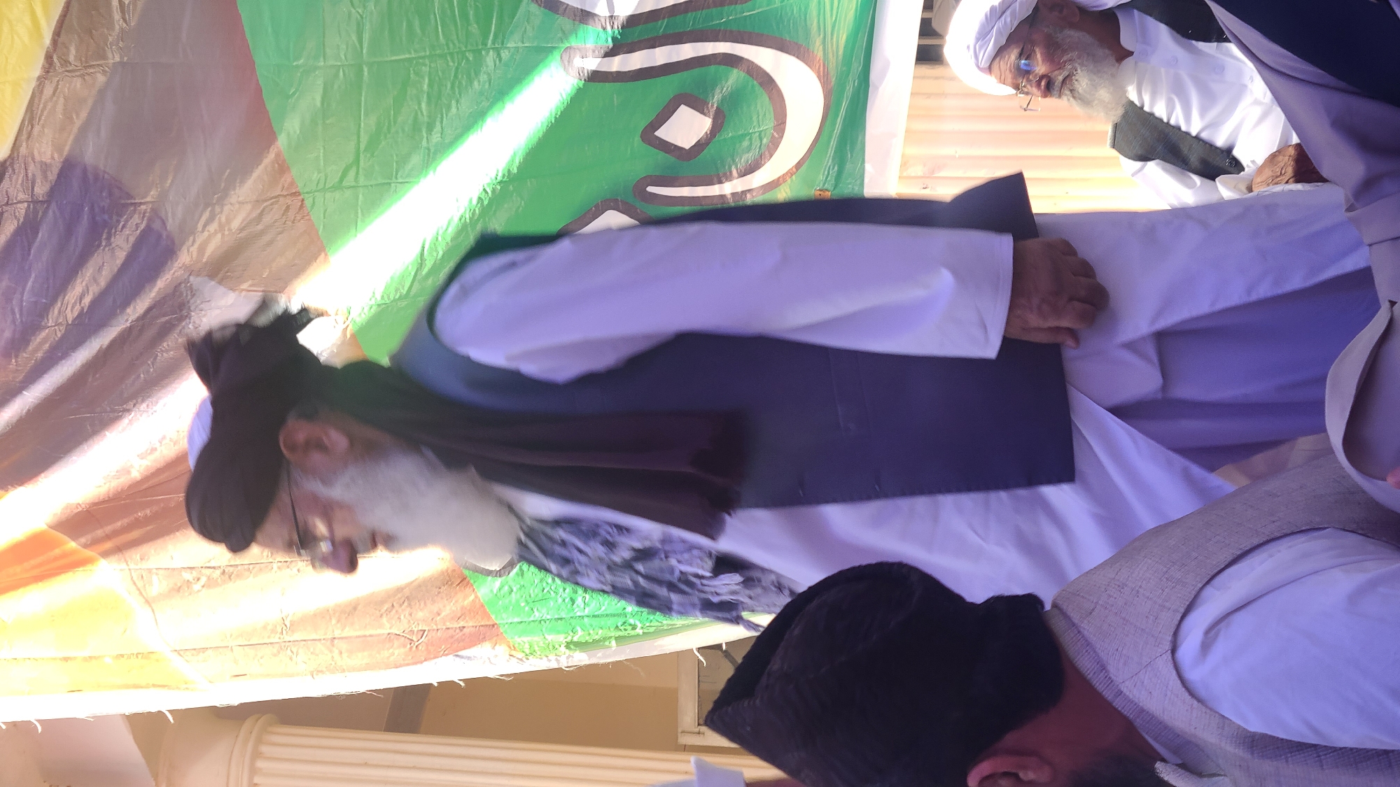 The image shows a group of men in Panjpai, Pakistan. Several men are wearing traditional Islamic attire, including white robes, black vests, and black hats. One man is wearing a white turban. The men are standing or sitting near a colorful banner that includes green and white colors. There is what appears to be a building support pillar in the lower left of the image.