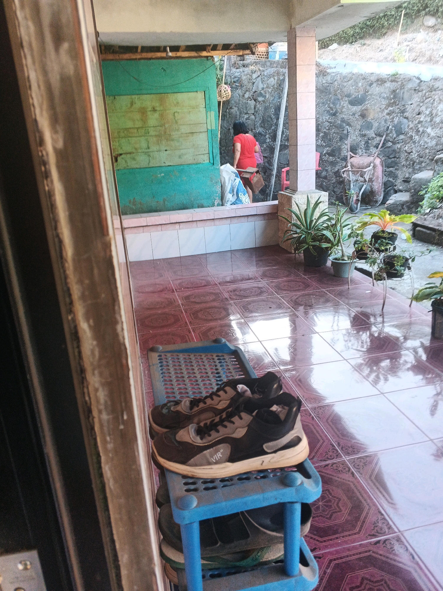 The image depicts a view from a doorway looking out onto a patio area. In the foreground, a blue plastic shoe rack is filled with shoes, with a pair of black and grey sneakers prominently displayed on the top shelf. The sneakers have "VIR" printed on the side. The patio floor is tiled with a reddish-pink patterned tile. Beyond the patio, there is a wall constructed from rough stones. A person wearing a red shirt is visible in the background, partially obscured by a column. To the right of the stone wall, a rusty wheelbarrow is present. Several potted plants are arranged on the patio. The environment suggests an outdoor residential setting, possibly in Keji, Indonesia, given the typical architectural and vegetation features.