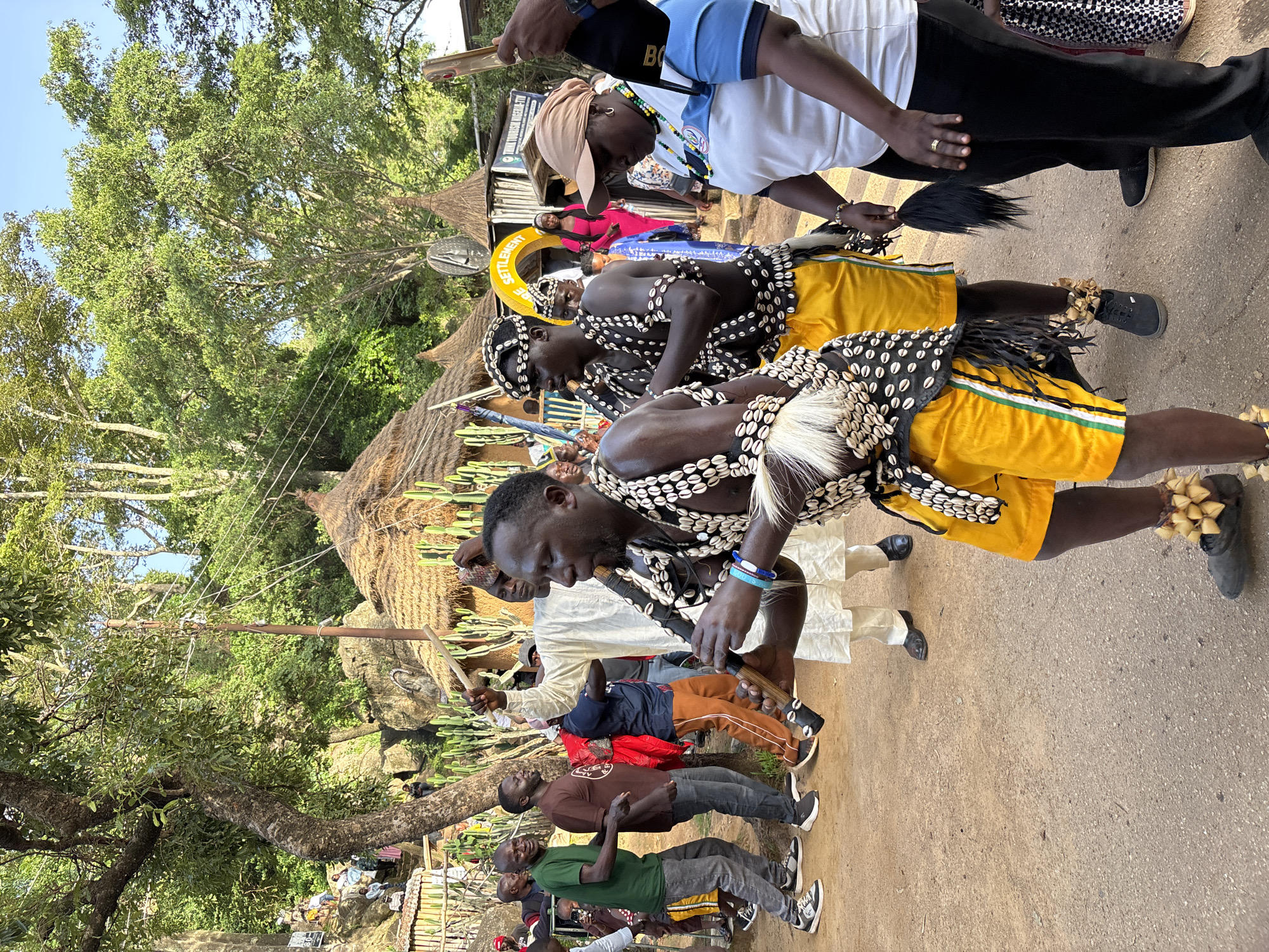 In Jos, Nigeria, a group of men are participating in a cultural event, possibly a festival or performance. The foreground is dominated by a man dressed in traditional attire, featuring a headdress adorned with shells, a necklace made of shells, and yellow shorts. He is playing a wooden flute-like instrument. To his left, another man in similar shell-adorned clothing is visible. Behind them, several other men are present. One man in a white robe is holding a stick aloft, while others are dressed in contemporary clothing. In the background, traditional thatched-roof huts are visible, suggesting a rural or traditional setting within the city. The environment appears to be outdoors, with trees and a clear sky. The overall scene depicts a lively cultural celebration.