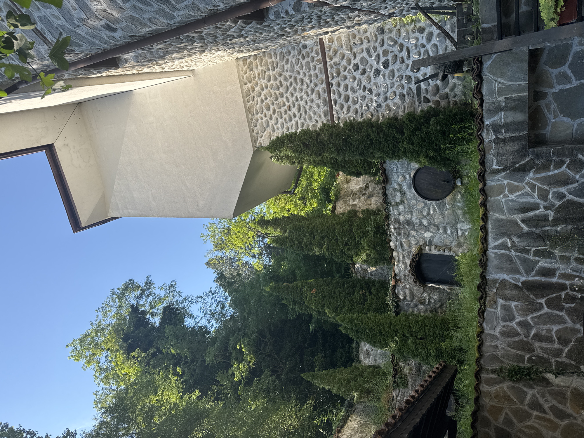 This image depicts an outdoor architectural and natural scene in Melnik, Bulgaria. The composition is dominated by multi-tiered stone walls and a modern building section, set against a backdrop of dense foliage and a clear blue sky.
In the upper right, a section of a white, smooth-textured building with sharp, angled lines and a prominent overhang is visible. A dark brown gutter runs along its upper edge. Below this modern structure, and extending to the right, a wall is constructed from numerous small, rounded, light grey river stones, mortared together. This wall incorporates horizontal bands of flat, darker grey stones. Below this, and continuing to the left across the middle ground, are retaining walls made of irregularly shaped, larger, flat grey stones with visible mortar lines. These lower walls are topped with rows of red-brown terracotta roof tiles, from which green grass and weeds grow.
Several tall, slender, dark green, conical evergreen trees are strategically planted on the terraces between the stone walls, providing vertical accents. On the far left, part of a dark wooden roof structure with terracotta tiles is visible. Integrated into one of the rounded-stone walls is a dark, arched wooden door. Further right on the same wall, a dark wooden barrel is horizontally affixed. On the far right, a segment of a rustic wooden staircase or railing, dark in color, descends.
The background, particularly on the left and center, features a steep hillside densely covered with lush, vibrant green deciduous trees and other foliage under a bright, clear blue sky. The scene is illuminated by natural sunlight, casting shadows that highlight the textures of the stone and the angularity of the white building. No people or animals are visible in the image.
