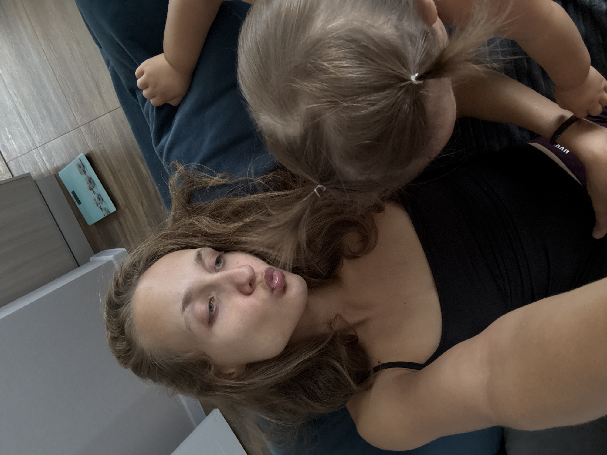 The image shows a close-up selfie of a young woman with her daughter. The woman is lying on a dark blue couch and is puckering her lips for the camera. Her daughter is leaning over her, and her hair is touching the woman's face. The daughter's hand is resting on the woman's arm. In the background, there is a digital scale on a tiled floor, and what appears to be a white cabinet. The overall scene suggests a casual, domestic setting, likely a home. There are no visible indicators of a specific city or country, so the location in Podshivalovo, Russia, cannot be confirmed from the image alone.