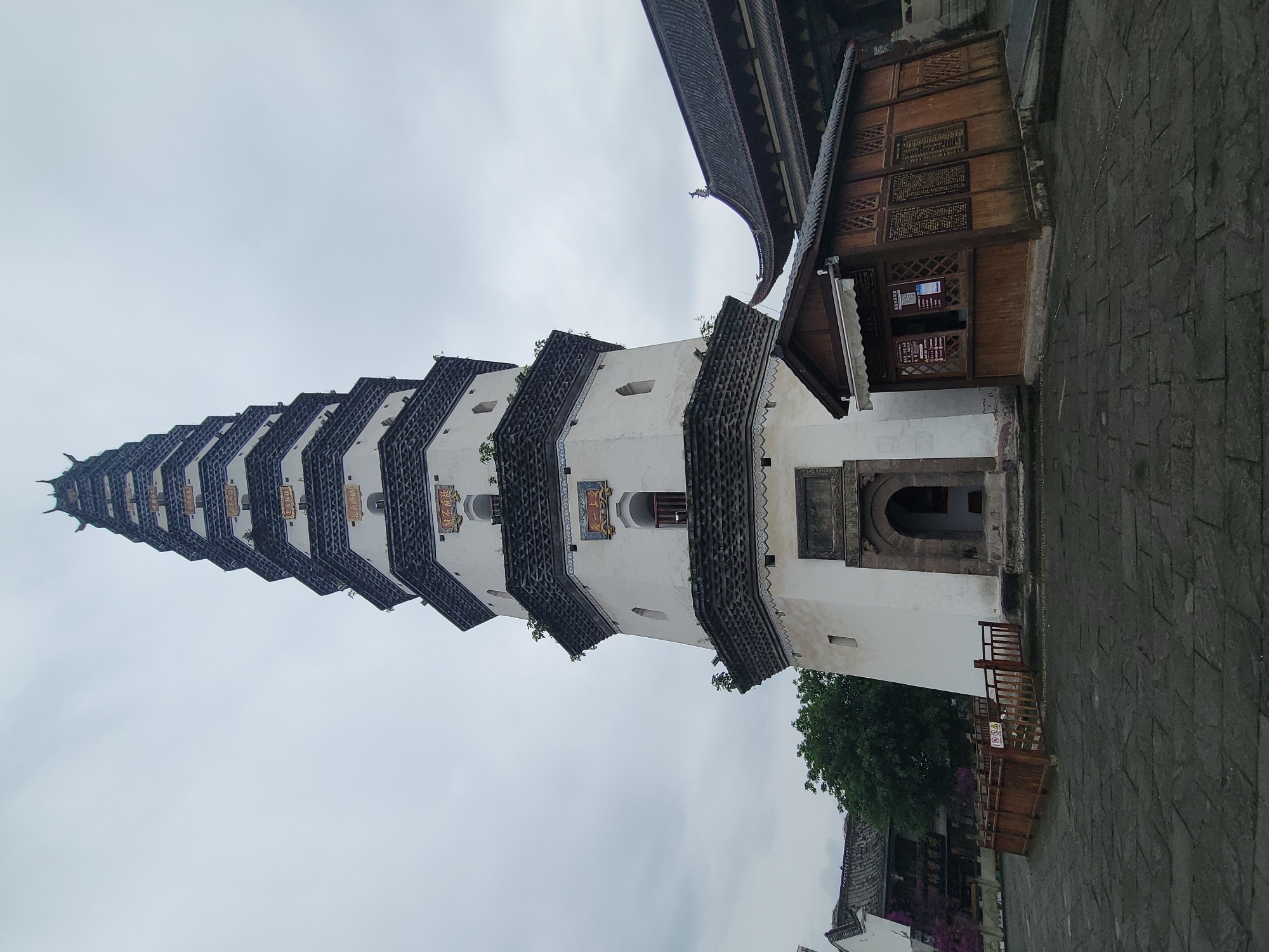 The image shows a tall pagoda-style building in Hechuan, China. The structure is white with dark gray tiered roofs. A smaller wooden building adjoins the base of the pagoda. The ground is paved with gray stones. The sky is overcast. A tree is visible near the base of the building. There are no people visible.