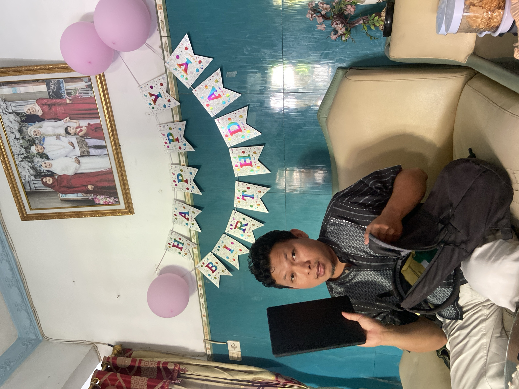The image captures an indoor scene, likely a living room or common area, with a man seated on a light-colored sofa. The background features a blue-green tiled wall and a white wall, adorned with birthday decorations.
The primary subject is a man with dark, curly hair, wearing a patterned, dark-colored short-sleeved shirt and light-colored trousers. He is looking directly at the camera while holding a black tablet or case in his left hand. An open black backpack rests next to him on the sofa, with some items visible inside.
Decorations indicate a birthday celebration: two pink balloons are visible on the left side, and a "HAPPY BIRTHDAY" banner hangs across the blue-green wall. The banner consists of individual flag-shaped pieces, each with a colorful letter.
Other objects in the scene include a large framed picture hanging on the white wall, depicting a group of several women and one child, some wearing traditional attire like kebaya. Text on the frame is partially visible, including "Kebaya." A small vase with artificial pink flowers is placed on the blue-green wall. On the right side, part of a clear jar filled with snacks or cereal is visible on a table next to the sofa. Red and gold patterned curtains can be seen in the lower left corner. The setting appears to be a cozy home environment.