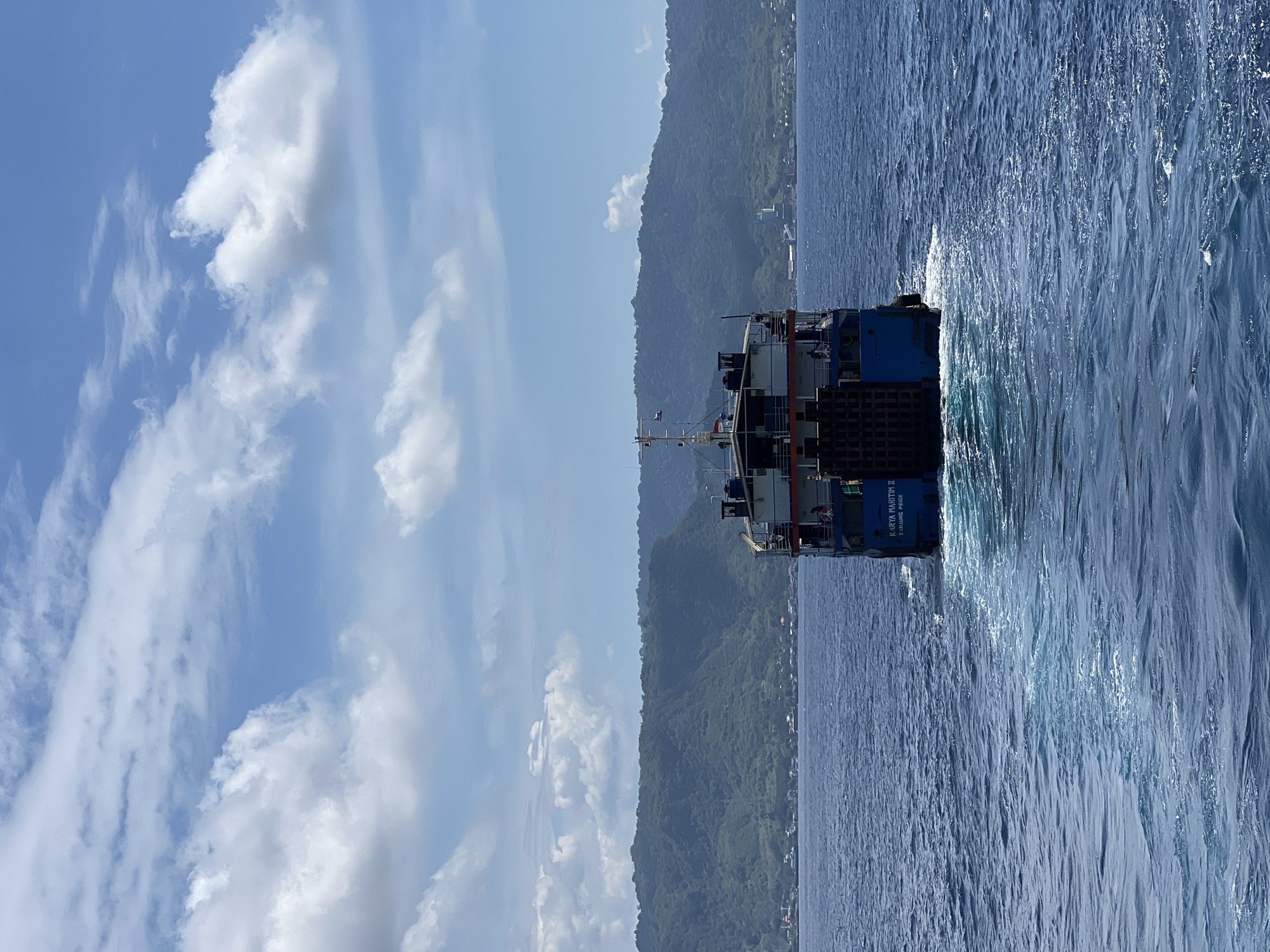 A medium-sized blue vessel is traveling on the ocean near a shoreline with a densely vegetated hill or mountain. The vessel has the name "Karya Maritim II Tanjung Priok" painted on its stern. The ocean waves indicate the ship is moving. The sky is blue with scattered white clouds. The shoreline is partially populated with structures visible amidst the vegetation. The specific city or location within Indonesia is not discernible from the image alone.