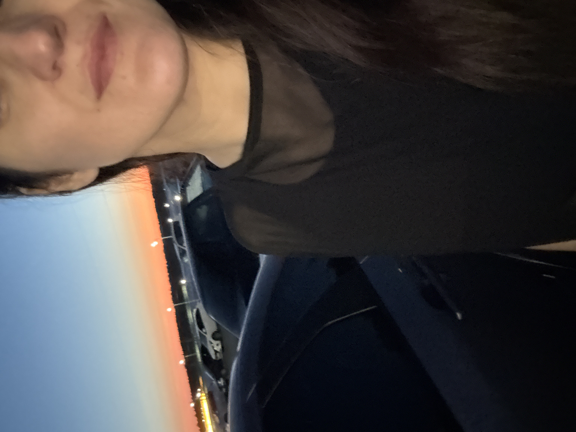 The image depicts a close-up of a woman's face and upper body, taken outdoors at dusk. She is wearing a black sheer top. In the background, a parking lot with several cars is visible, and a sunset with orange and blue hues streaks across the sky. The setting appears to be a city street in Sarapul, Russia, indicated by streetlights and the overall urban landscape. The lighting suggests it is evening.
