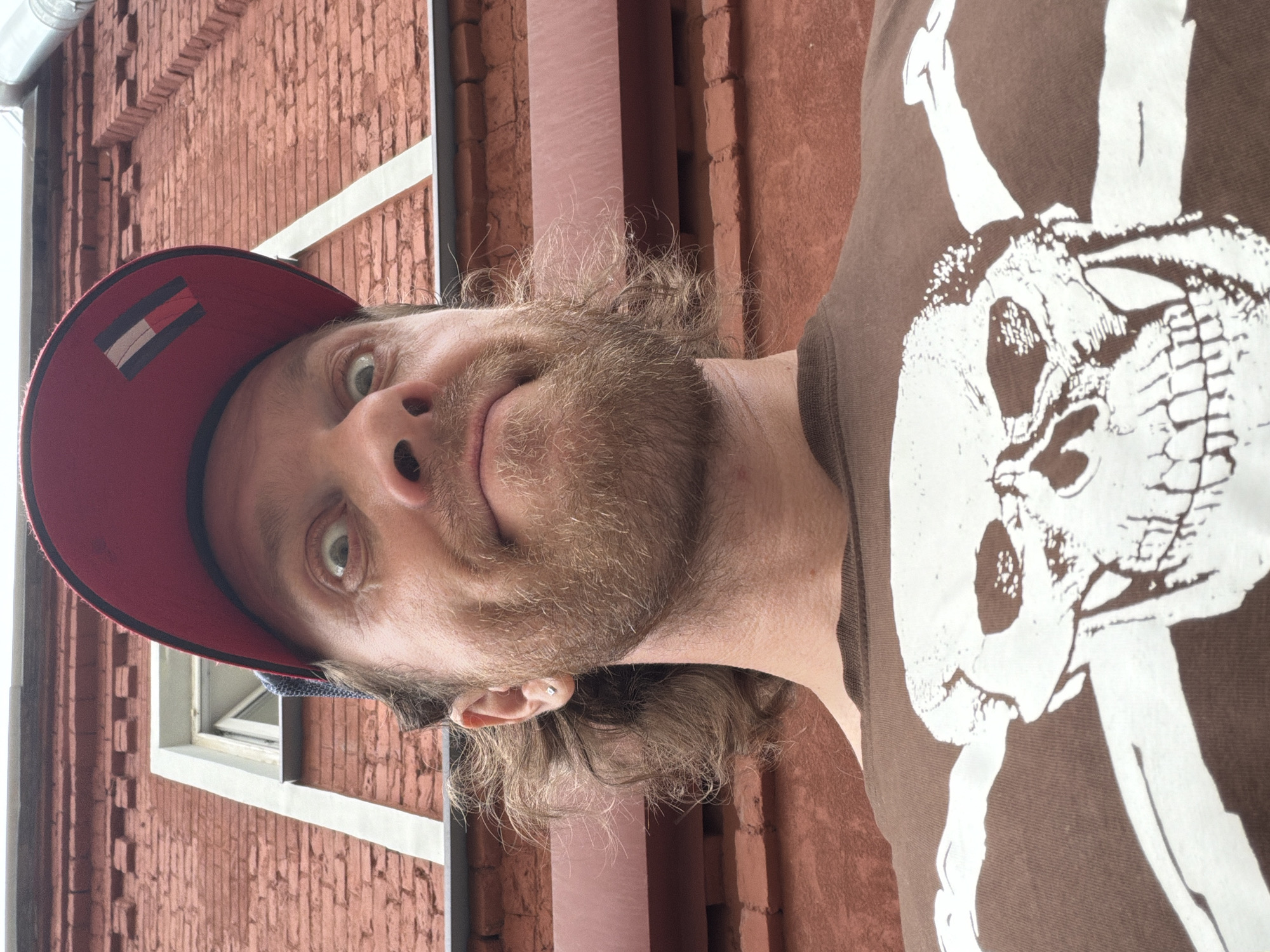A low-angle, close-up, point-of-view shot captures a man wearing a red baseball cap and a brown t-shirt with a white skull and crossbones graphic. The man has light brown hair, a beard, and is looking upwards with a surprised expression, his eyes wide open. He has a small silver earring in his left ear. The background consists of a red brick building with a white trim around a window and a dark red metal awning. The building appears to be an exterior wall of a structure located in the city of Saint Petersburg, Russia.