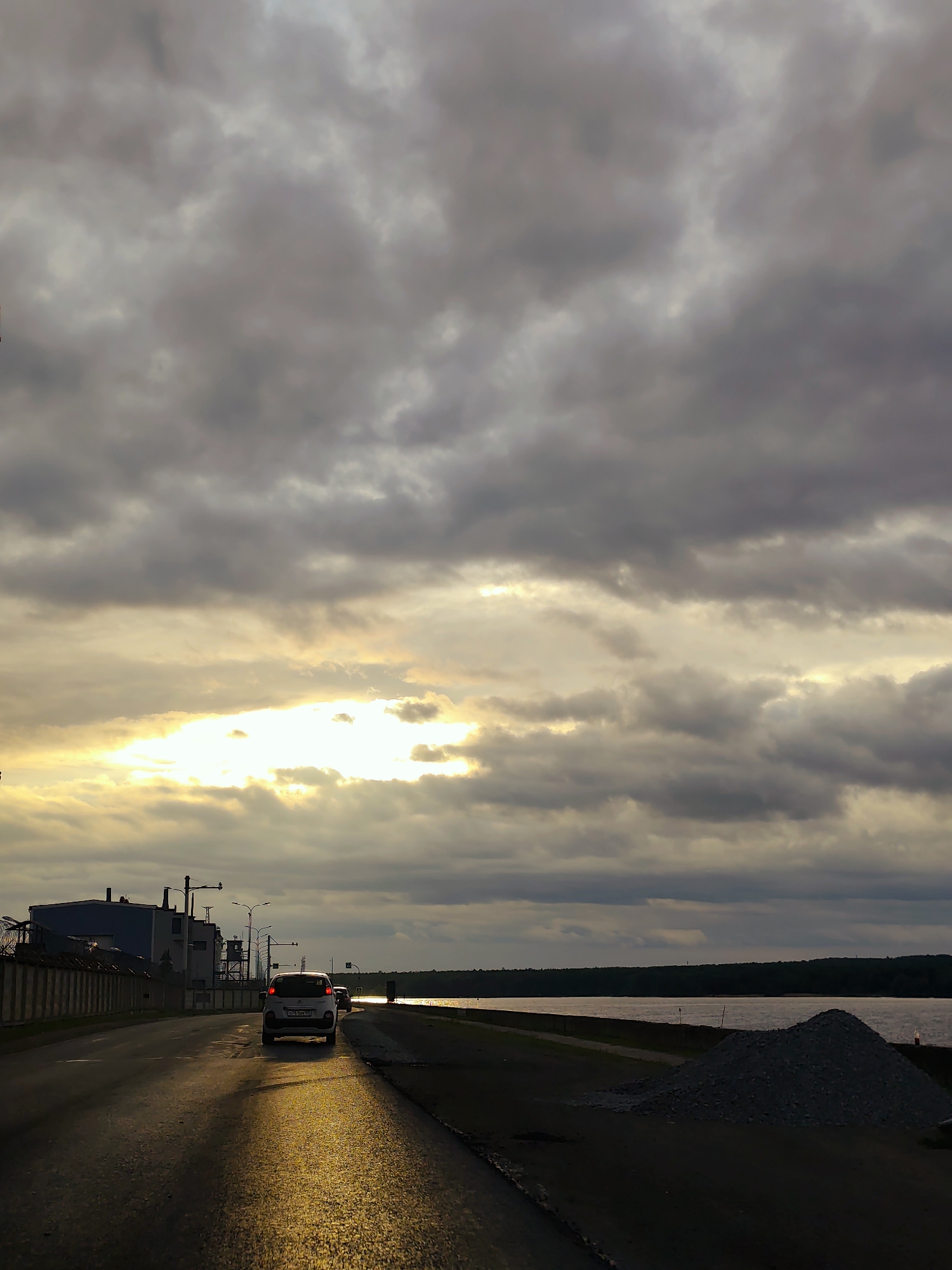 This image captures a scene along a paved road in Chaikovsky, Russia, at a time of day consistent with late afternoon or early evening, characterized by an overcast sky with a distinct break through which bright sunlight penetrates. This sunlight creates a prominent golden reflection on the road surface in the foreground and midground.
In the mid-ground, a white compact car, identified by its illuminated red rear lights, is traveling away from the viewer. A portion of its Russian license plate is visible, displaying "O 256 HO 18". Another vehicle is partially obscured behind the white car. To the immediate right of the road, a large, conical pile of light grey aggregate material, likely gravel or crushed stone, rests on an unpaved shoulder, indicating potential construction or roadwork.
On the left side of the road, a concrete barrier runs parallel to the asphalt. Beyond this barrier, industrial structures with white and blue elements, along with various pipes and fixtures, are visible. A fence topped with barbed wire is also present, separating the road from these facilities.
To the right, beyond the aggregate pile, a wide body of water, consistent with the Votkinsk Reservoir on the Kama River, stretches into the distance. The water's surface reflects the sunlight, creating a bright band near the horizon. The far bank of the body of water is marked by a dense line of dark green trees. The sky above is largely covered by dark grey and white clouds, with the intense light source emanating from a clear patch in the cloud cover.