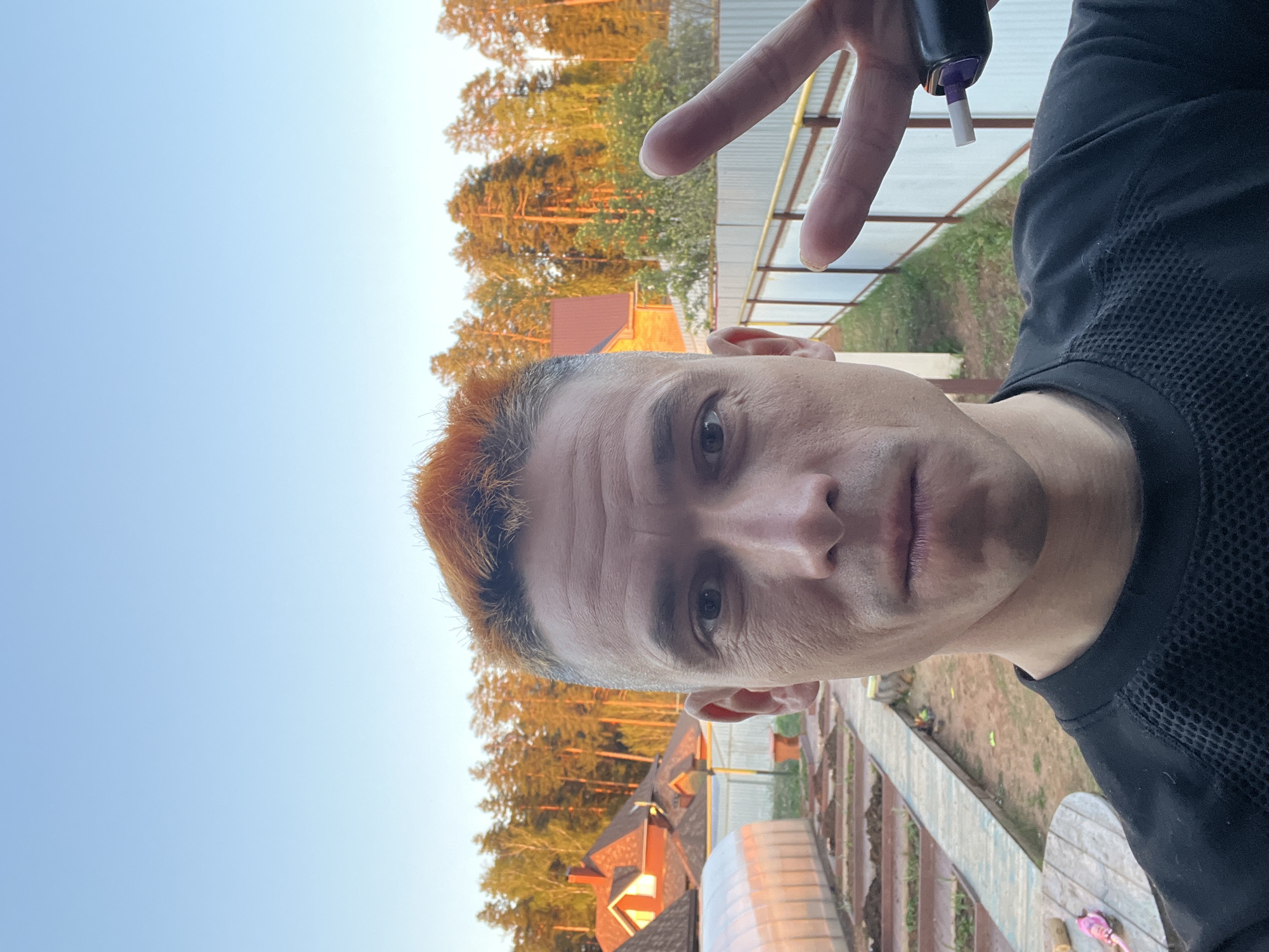 A close-up, tilted shot of a man with dyed, orange-tinted hair standing outside in what appears to be a rural or suburban setting. He is wearing a black shirt with a mesh-like texture around the collar. He holds what appears to be a vape pen in his hand. Behind him is a background that includes trees, houses with visible roofs, and a small greenhouse.
While the location cannot be definitively confirmed as Chaikovsky, Russia, the presence of what could be typical Russian-style houses and vegetation does not contradict this assertion. The photo appears to have been taken outdoors, with natural lighting.