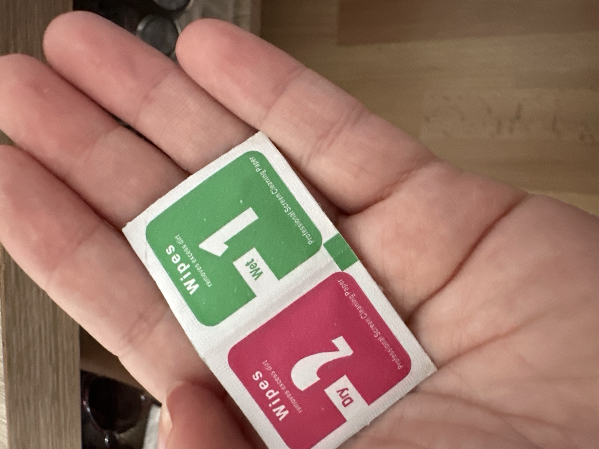 A person's hand is holding a package of two screen cleaning wipes. One wipe is pink and labeled "Dry," and the other is green and labeled "Wet." The wipes are designed for professional screen cleaning. The background appears to be a wooden surface. The location is not specified in the image or the provided crops.