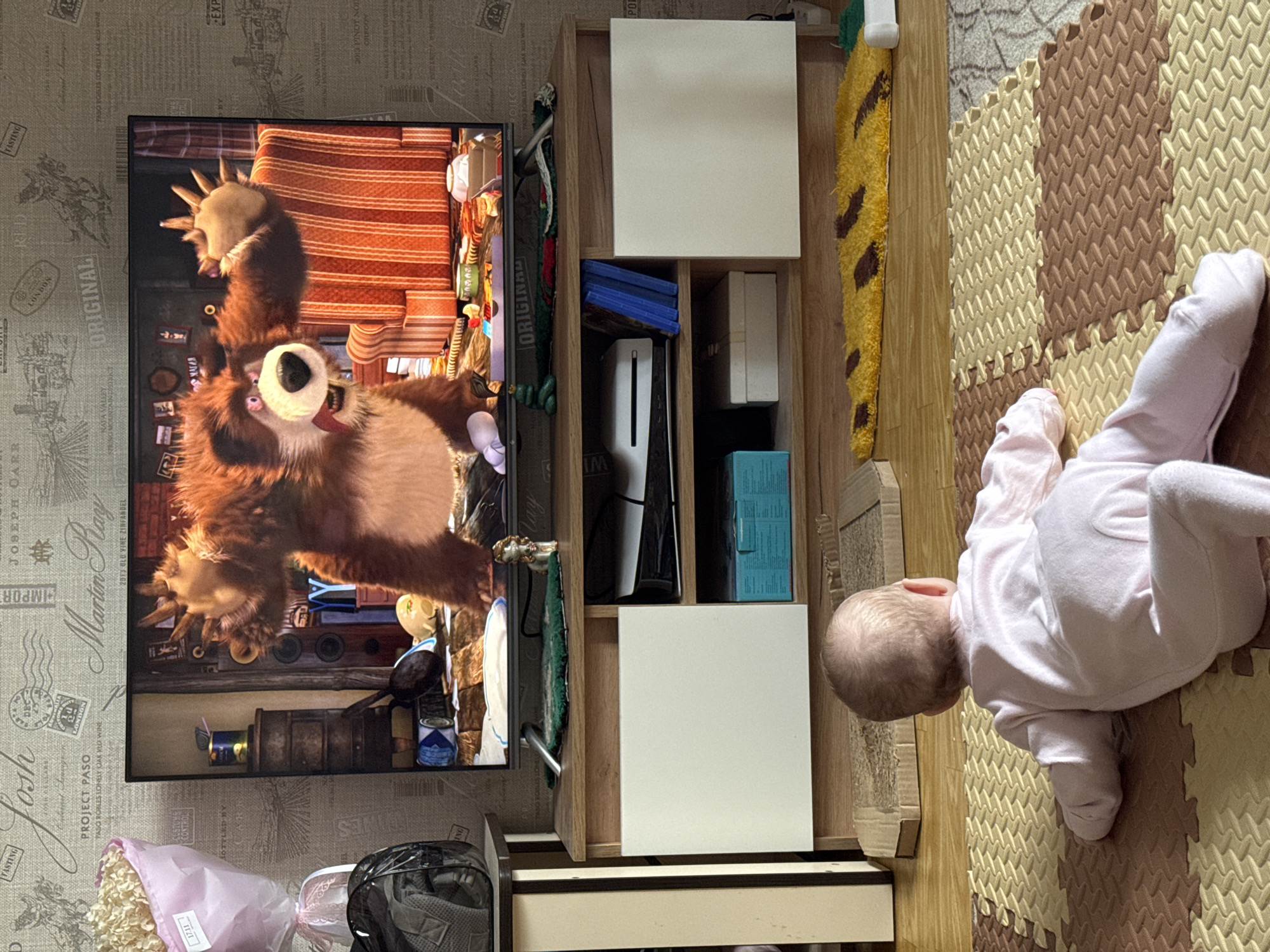 A domestic scene shows a baby lying on a foam play mat next to a wooden shelf unit with assorted items including what looks like a PlayStation 5 console. On the wall to the left is a television playing the Russian animated series "Masha and the Bear." The photo appears to be taken indoors, in a room with patterned wallpaper. Some flowers in pink wrapping are placed at the bottom left corner of the image. Location is inferred to be the city of Chaikovsky, Russia.