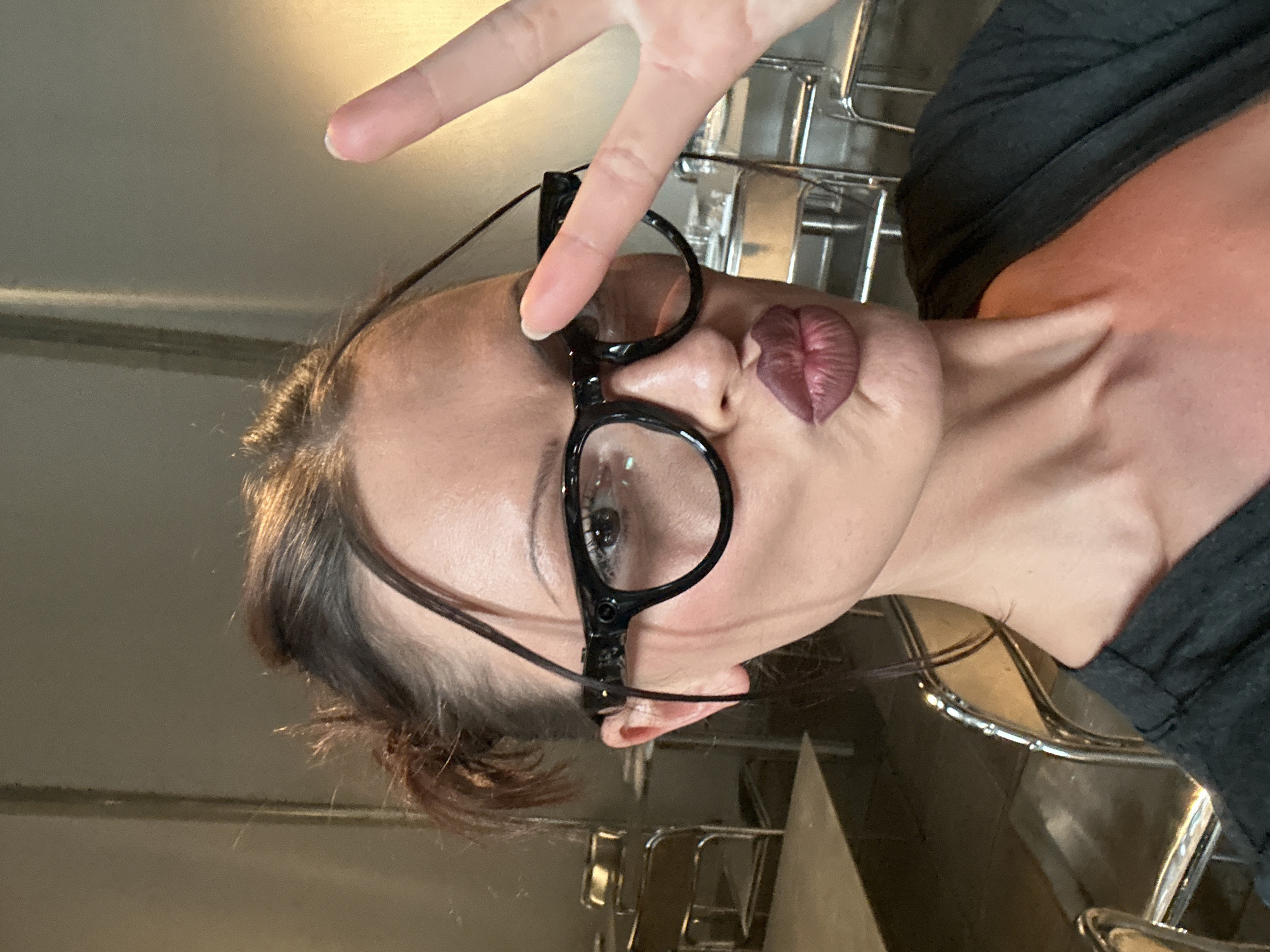 A woman with brown hair pulled back in a bun is indoors and angled away from the camera, holding up a peace sign that obscures the glasses she is wearing. She is wearing a dark colored shirt. Her lips are shaped like a heart. The background consists of walls and chairs, possibly made of metal. The location in London, UK, is unknown.