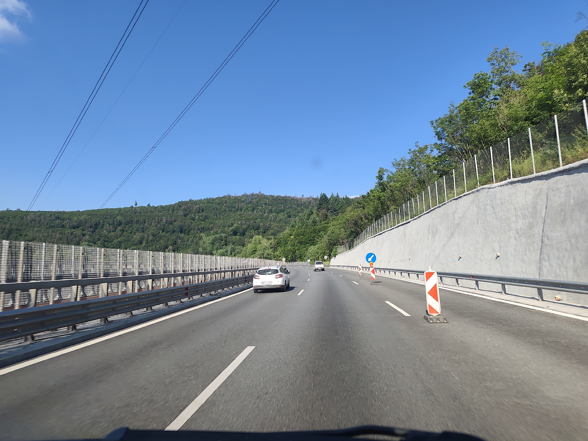 This image captures a view from a vehicle driving on a multi-lane highway through a mountainous, forested area. The highway is paved with asphalt and marked with white lines. On the left side of the road, there is a metal guardrail and a sound barrier made of metal mesh and concrete panels. On the right side, a grey concrete retaining wall slopes upwards, topped with a metal fence. Trees and dense green foliage cover the hillsides in the background. Several vehicles are visible on the highway, including a white car in the foreground and another car further ahead. Traffic cones, including a distinctive orange and white striped marker and a blue directional sign, are present on the right side of the road, suggesting a lane closure or construction. Overhead, power lines stretch across the clear blue sky. The scene appears to be on the Hemus Motorway (A2) near Vrachesh, Bulgaria, a route known for its scenic mountainous terrain.