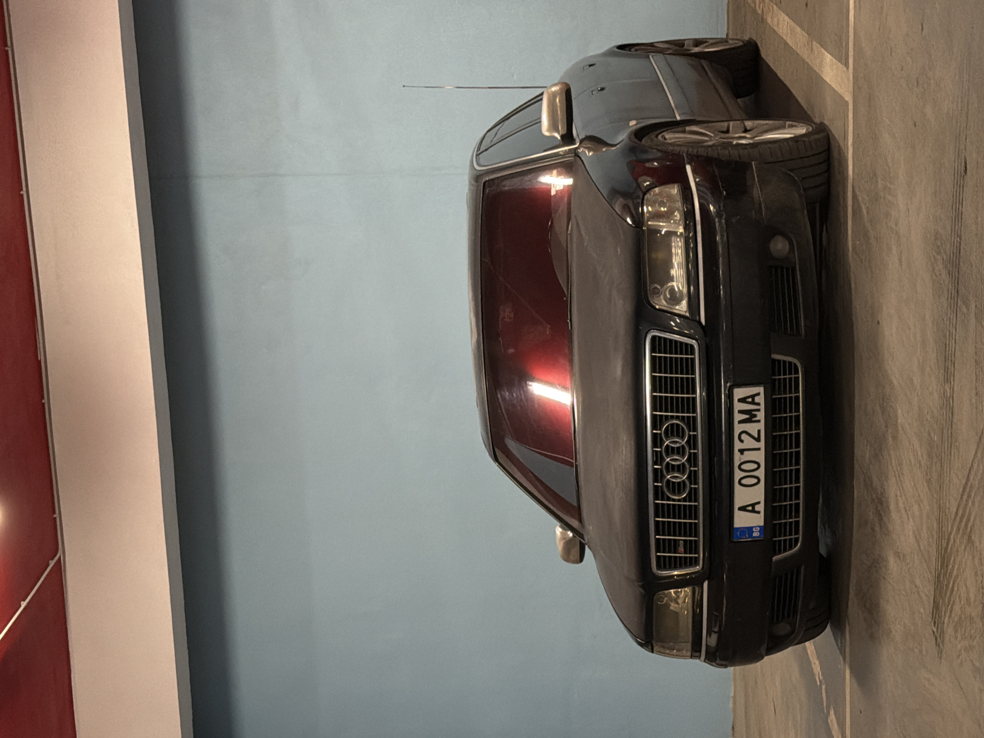 A medium shot shows a black Audi passenger car parked in what appears to be an indoor parking facility. The car is positioned at an angle within its parking space. It has the Bulgarian license plate "A 0012 MA." The background consists of a pale blue wall on one side and a grey concrete floor on the other. There are no visible people or indications of the car's location within Varna, Bulgaria, other than the license plate. The lighting is artificial.
