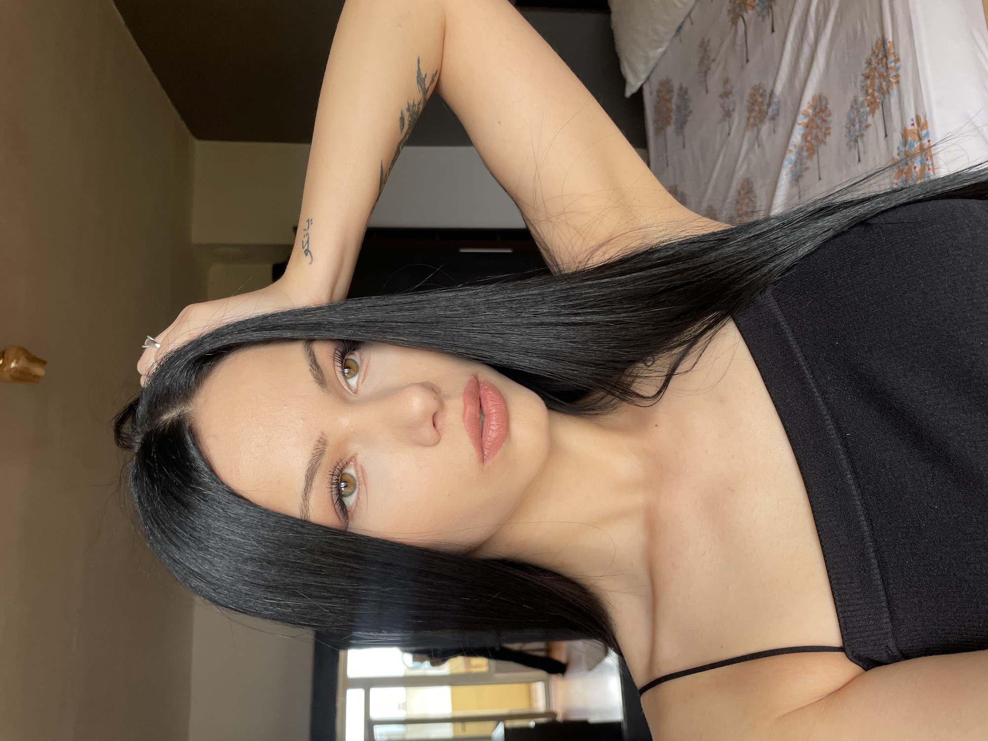 The image shows a close-up of a woman with long, straight black hair and light brown eyes. She is wearing a black tank top with thin straps. Her left hand is raised to her head, and she is wearing a ring on her left index finger. There is a tattoo on her left forearm. The background is slightly blurred but appears to be an indoor setting, possibly a bedroom. A bed with a patterned duvet is visible on the right side of the image. On the left side, a doorway with a person standing in it is visible through a window or open door. The overall lighting suggests it is daytime. The image does not contain any specific landmarks or details that would allow for its location to be identified as Stara Zagora, Bulgaria.